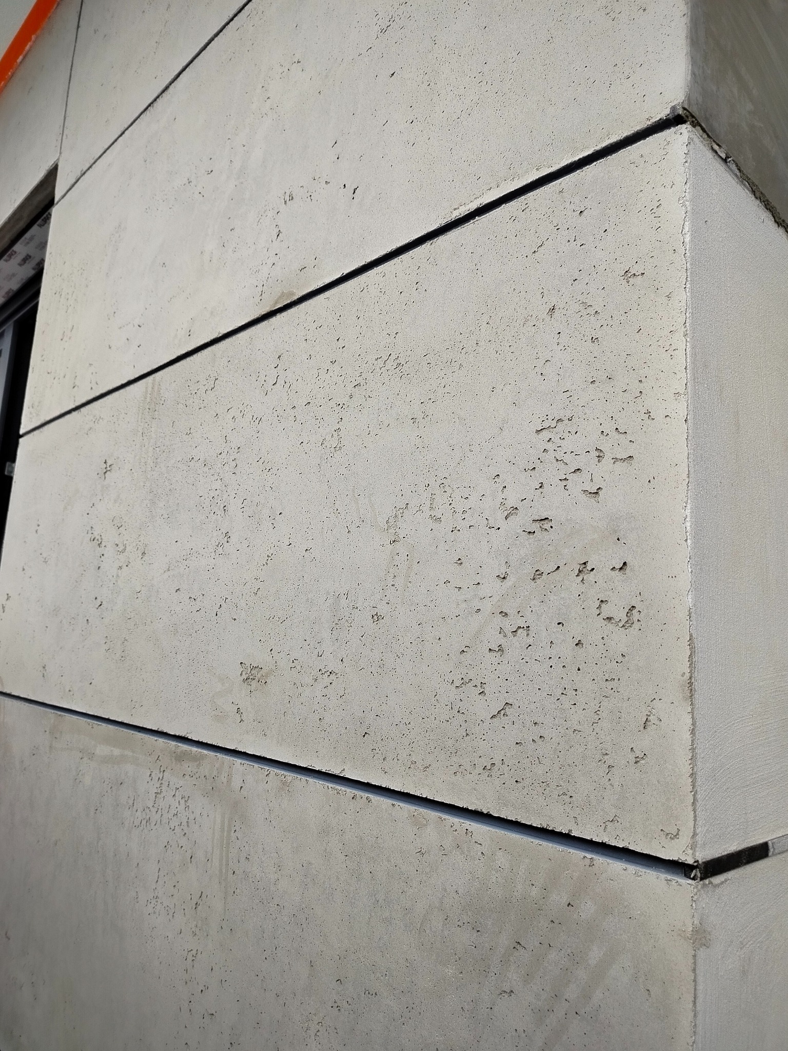 Szara, segmentowa elewacja budynku z wyraźną fakturą, widoczna z bliska pod kątem. Ciemne, poziome szczeliny między segmentami. Fragment okna po lewej stronie.