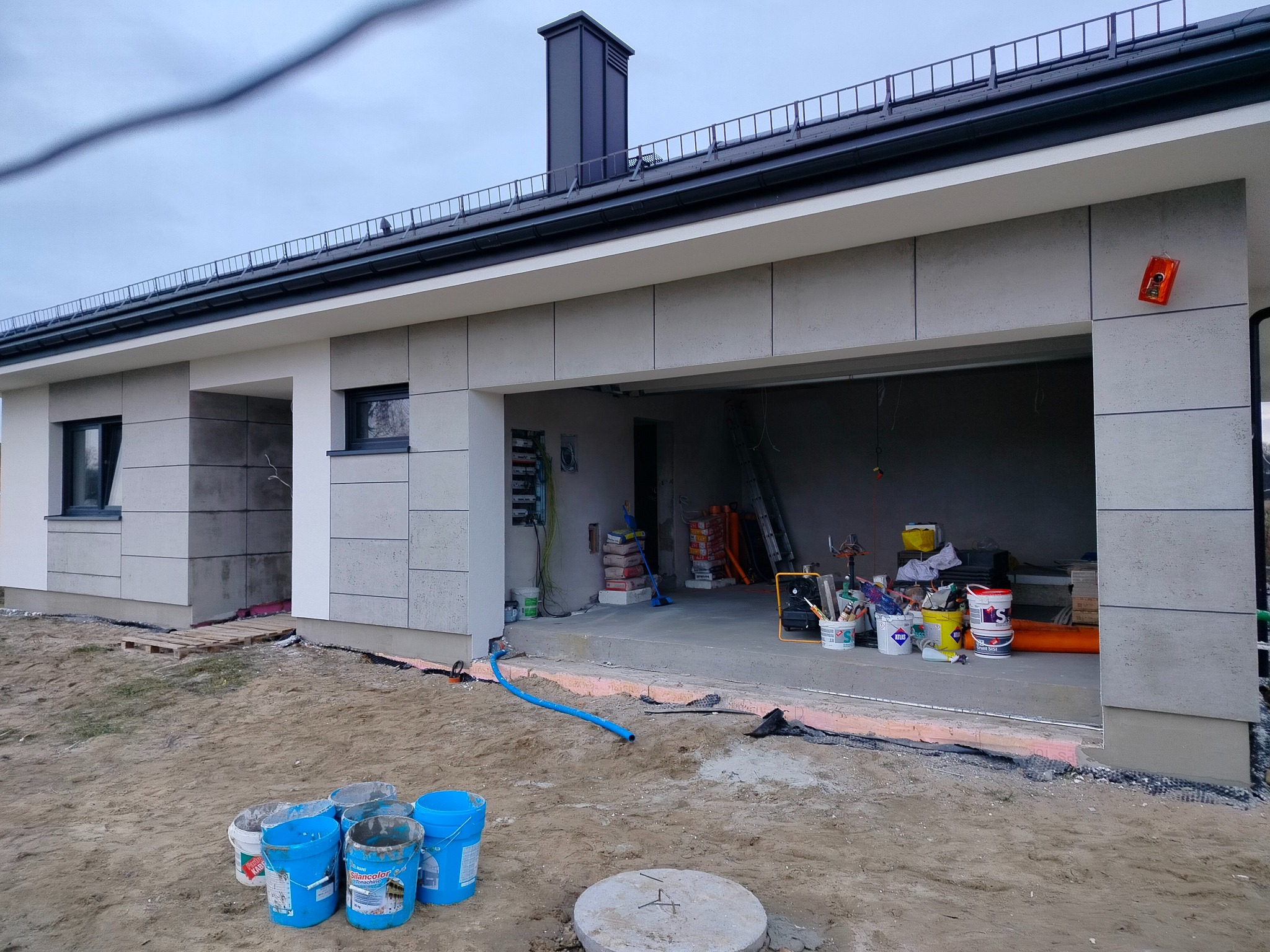 Nowoczesny dom jednorodzinny w trakcie wykończenia elewacji, widoczne wiaderka z farbą i narzędzia. Częściowo otynkowana ściana, surowy teren wokół budynku.