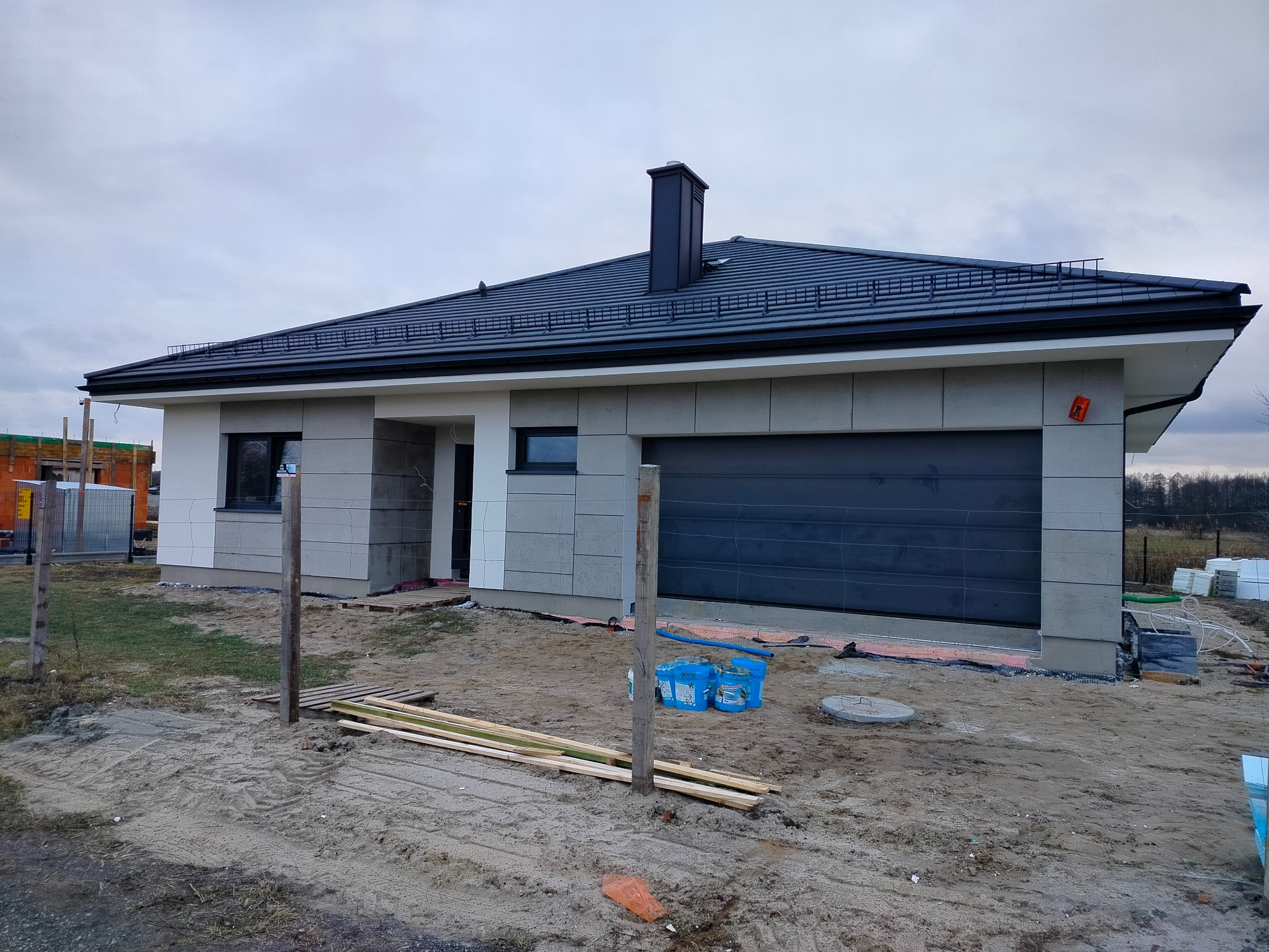 Nowoczesny dom jednorodzinny z grafitową bramą garażową i jasną elewacją w trakcie wykończenia. Widoczne materiały budowlane i elementy krajobrazu.