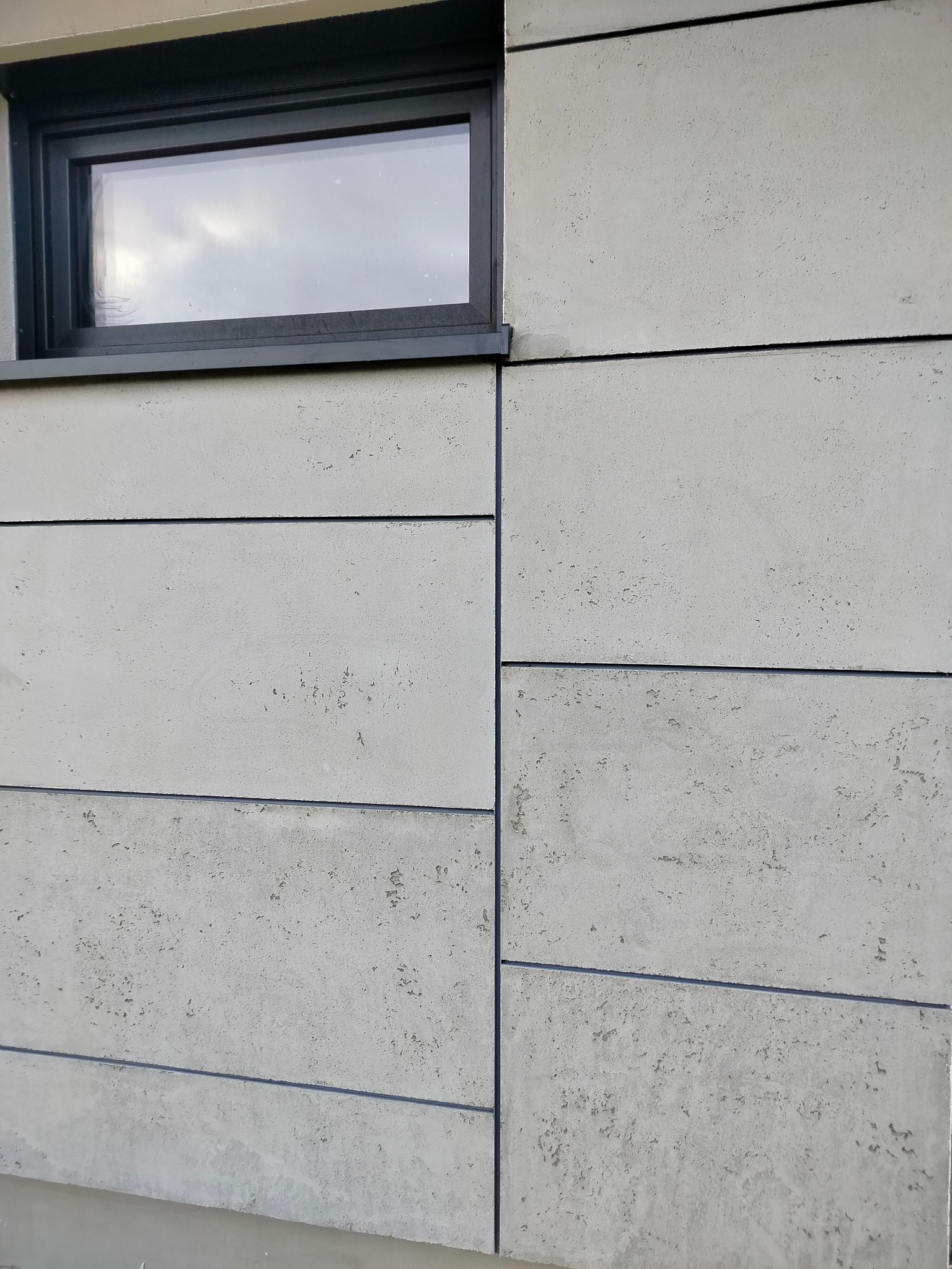 Szara elewacja budynku z poziomymi panelami i oknem z czarną ramą. Widoczna faktura paneli i precyzyjne łączenia. Minimalistyczny design.