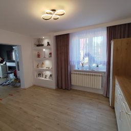 Szymon Zasępa - Jasne wnętrze po remoncie: drewniana podłoga, białe ściany z półkami, okno z zasłonami i grzejnikiem. Widok na salon z kominkiem. Nowoczesne oświetlenie sufitowe.