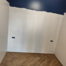 Szymon Zasępa - Ściana w trakcie malowania na biało i granatowo, widoczne gniazdka elektryczne i parkiet w jodełkę. Częściowo pomalowany sufit. Etap prac wykończeniowych.