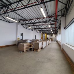 Szymon Zasępa - Magazyn z paletami płyt gipsowo-kartonowych i krzesłem. Widoczne białe ściany, metalowa konstrukcja dachu i okna z białymi lamelami. Perspektywa z głębi magazynu.