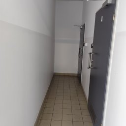 Szymon Zasępa - Korytarz z dwiema szarymi drzwiami z metalowymi klamkami i systemem samozamykającym. Podłoga wyłożona kwadratowymi płytkami w odcieniach beżu. Ściany białe z szarym pasem.