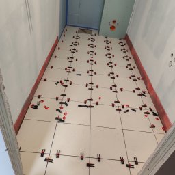 Szymon Zasępa - Białe płytki podłogowe w trakcie układania w wąskiej łazience. Czerwone klipsy poziomujące. Ściany przygotowane pod płytki, częściowo z płyt karton-gips.