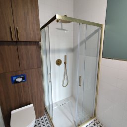 Szymon Zasępa - Nowoczesna kabina prysznicowa z złotymi profilami, białe płytki i wzorzysta podłoga w łazience. Umywalka podwieszana i szafka z drewnianymi frontami.