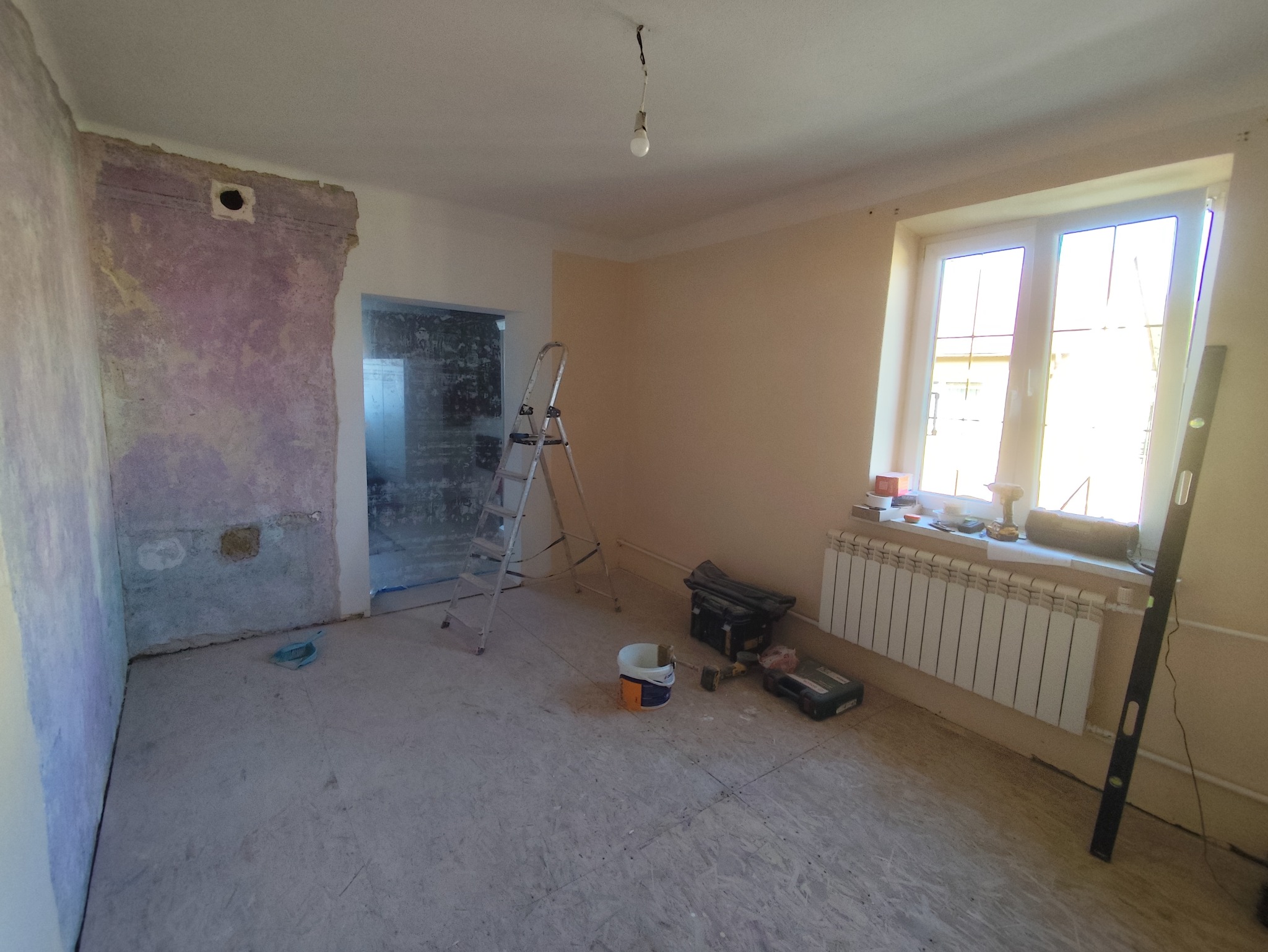 Pokój w trakcie remontu: ściany częściowo otynkowane, drabina, narzędzia malarskie, okno z grzejnikiem. Widoczne przygotowania do malowania lub szpachlowania.
