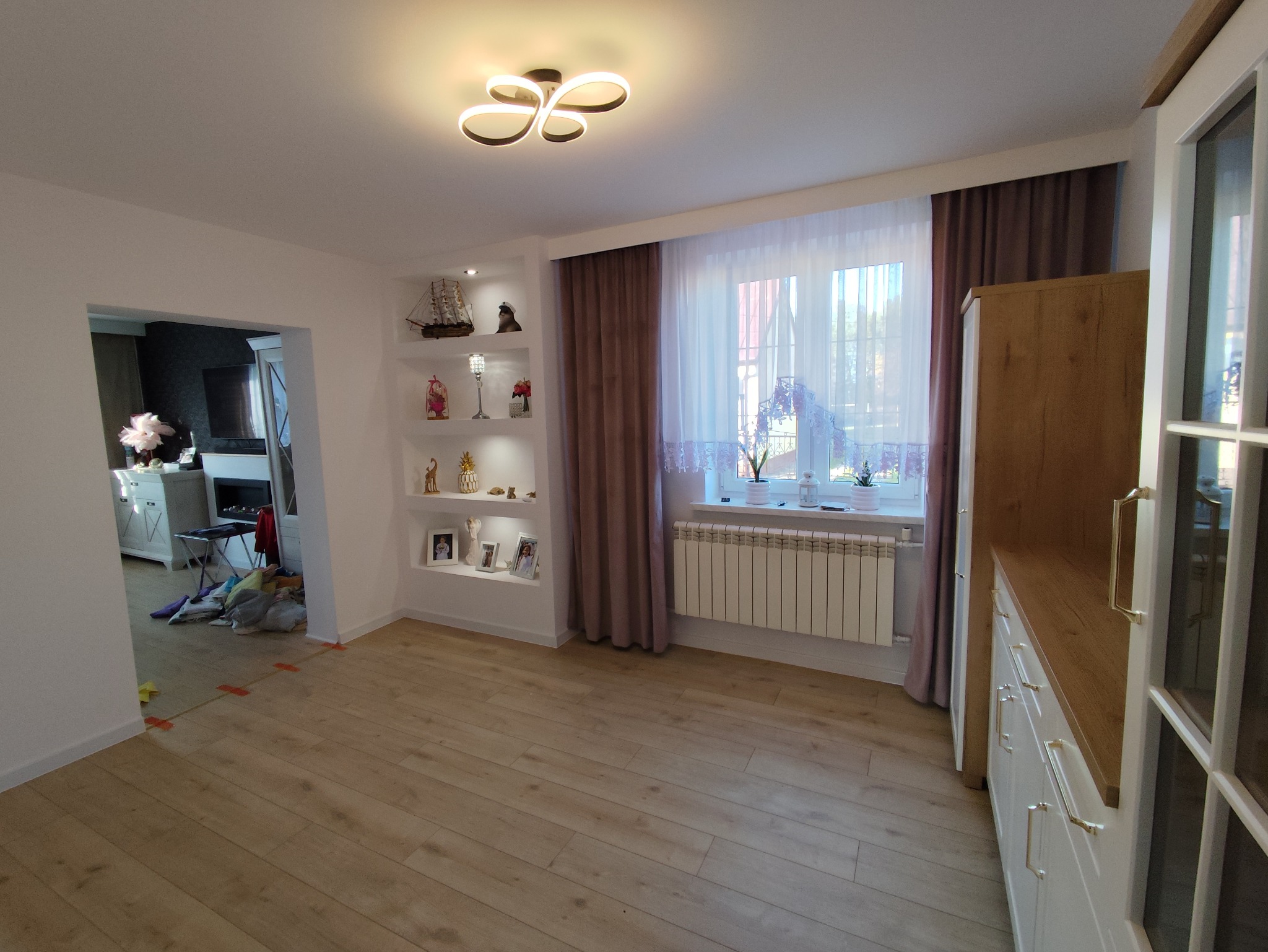 Jasne wnętrze po remoncie: drewniana podłoga, białe ściany z półkami, okno z zasłonami i grzejnikiem. Widok na salon z kominkiem. Nowoczesne oświetlenie sufitowe.