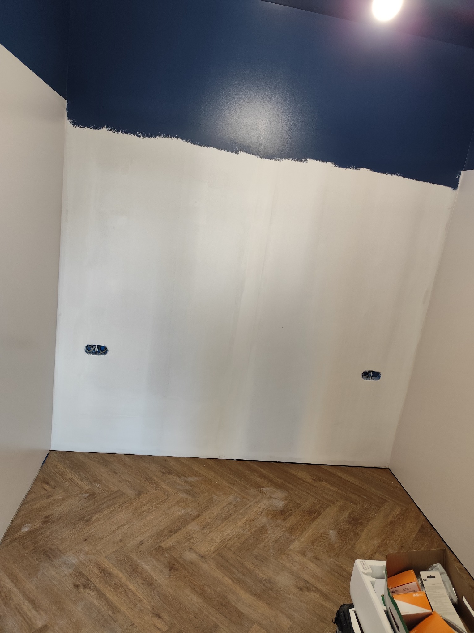 Ściana w trakcie malowania na biało i granatowo, widoczne gniazdka elektryczne i parkiet w jodełkę. Częściowo pomalowany sufit. Etap prac wykończeniowych.