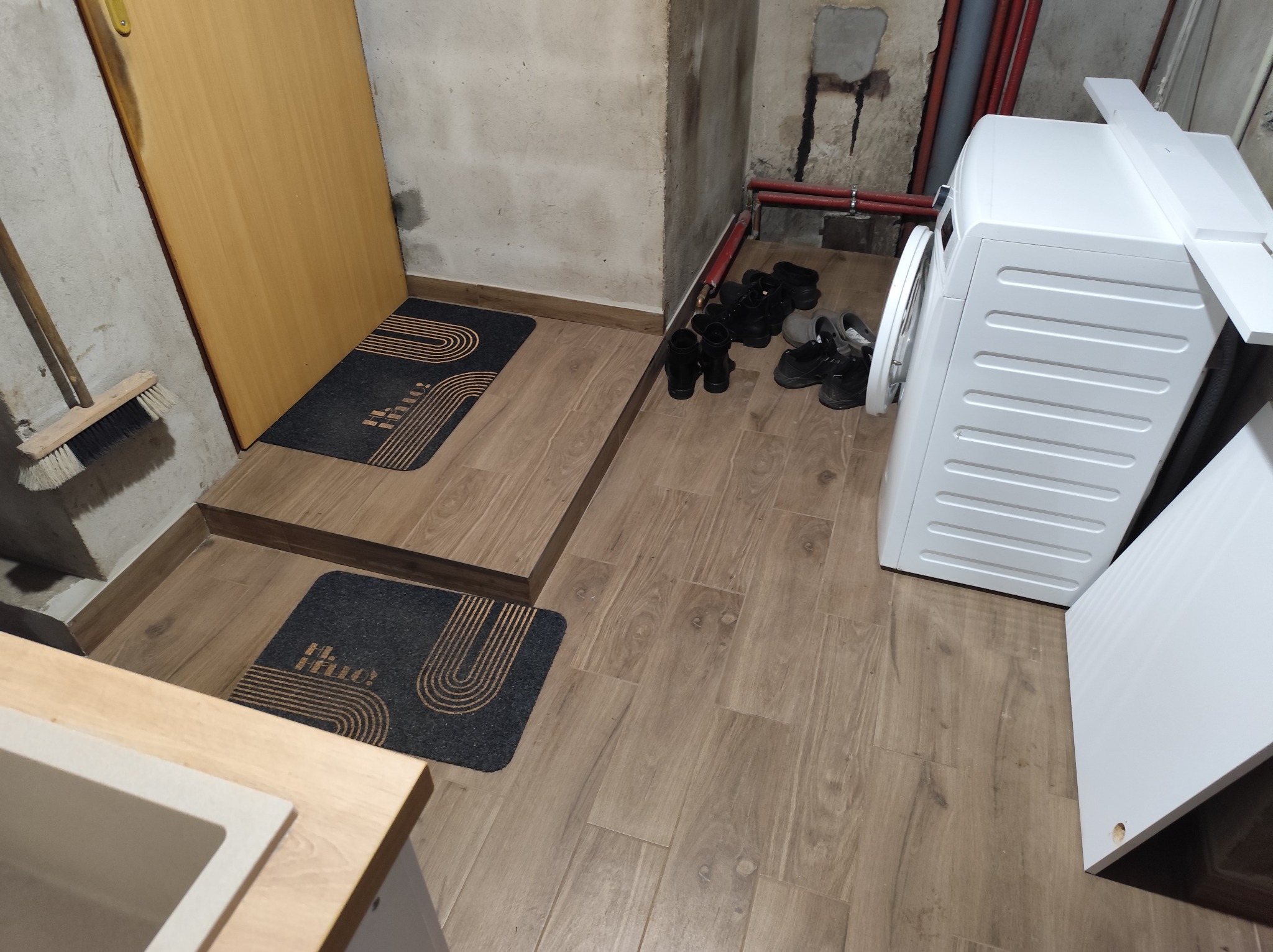 Ukończona podłoga z paneli drewnopodobnych w piwnicy, z widoczną pralką, drzwiami i dywanikami. Widoczne rury i buty.