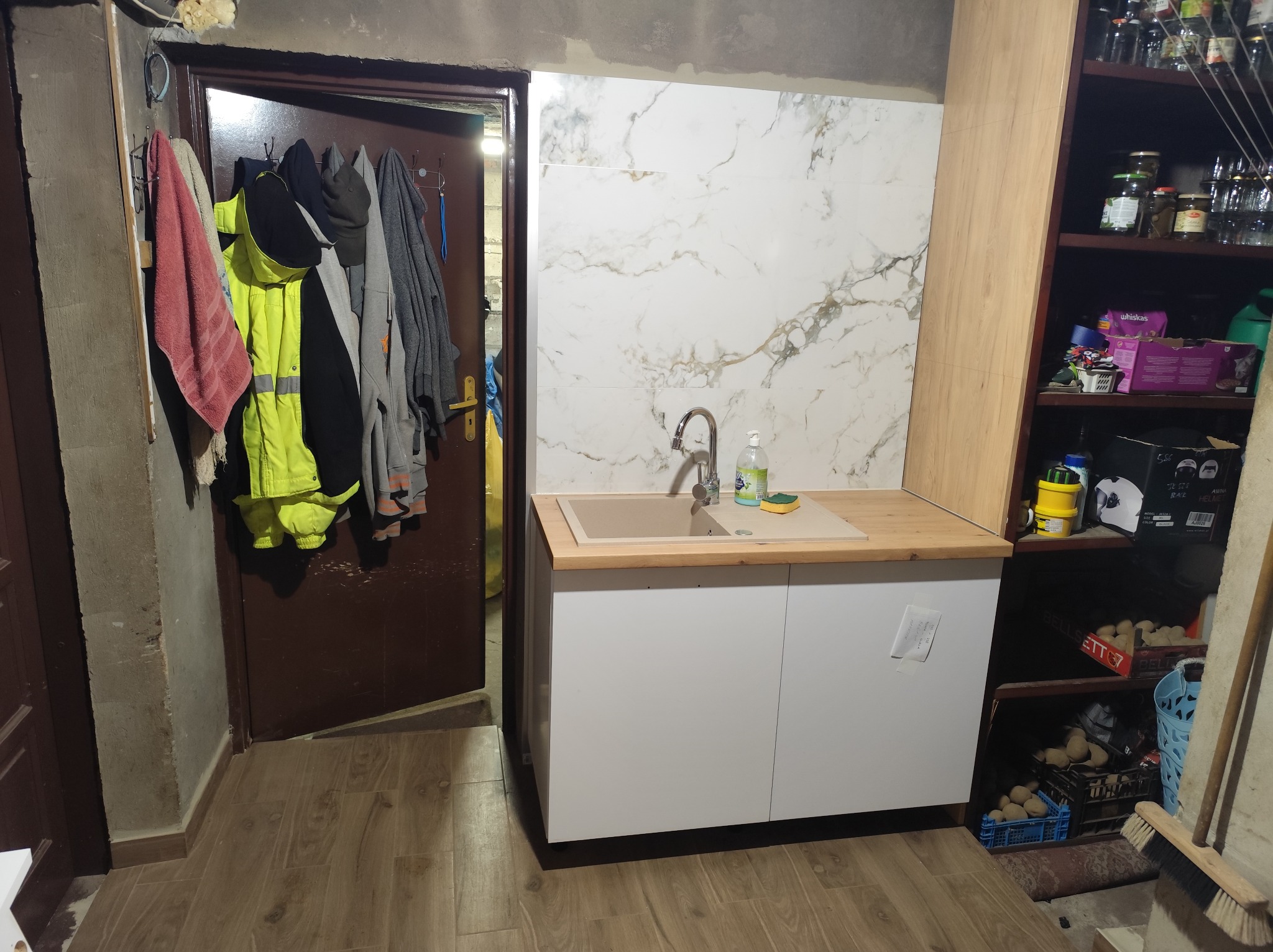 Wykończona strefa zlewu w kuchni: białe szafki, drewniany blat, marmurowe płytki na ścianie. W tle widoczne przedmioty codziennego użytku.