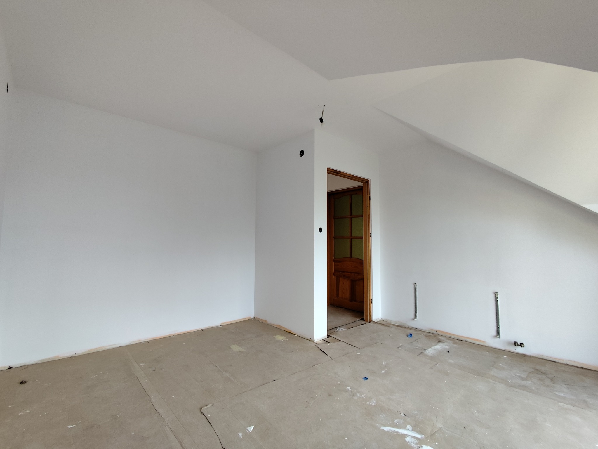 Wnętrze pokoju na poddaszu po malowaniu ścian na biało. Widoczne gładkie ściany, drewniane drzwi i przygotowane punkty elektryczne. Podłoga wymaga wykończenia.