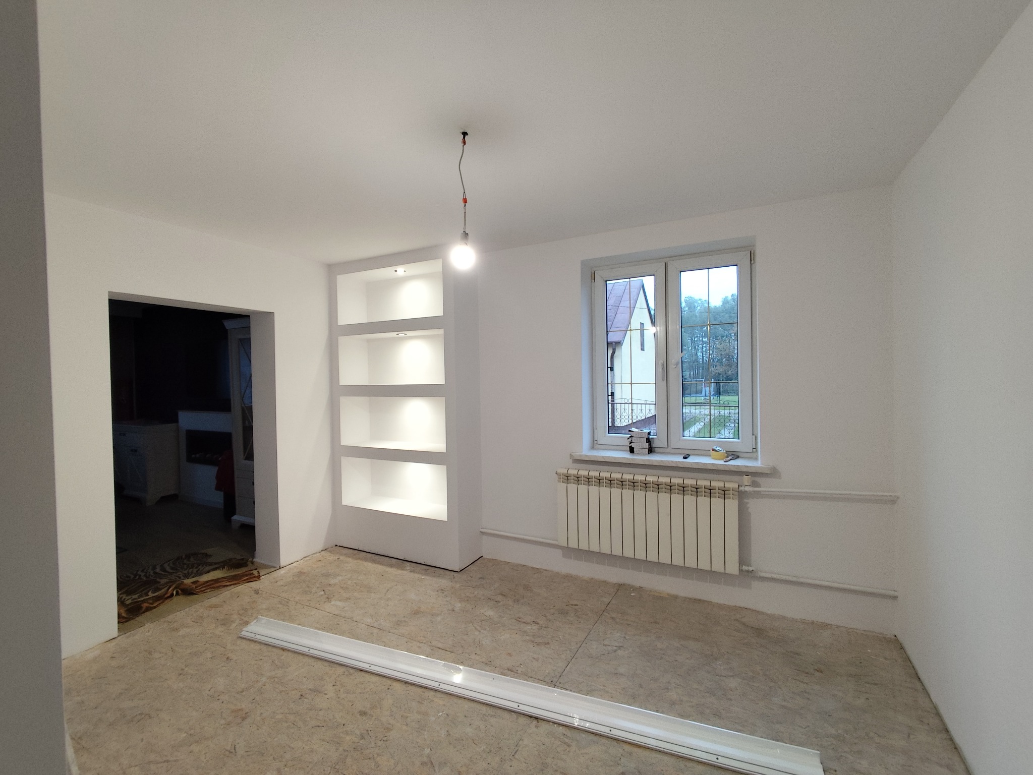 Białe wnętrze po remoncie: wnęka z półkami z oświetleniem, okno z grzejnikiem, surowa podłoga. Widok na sąsiedni dom przez okno.