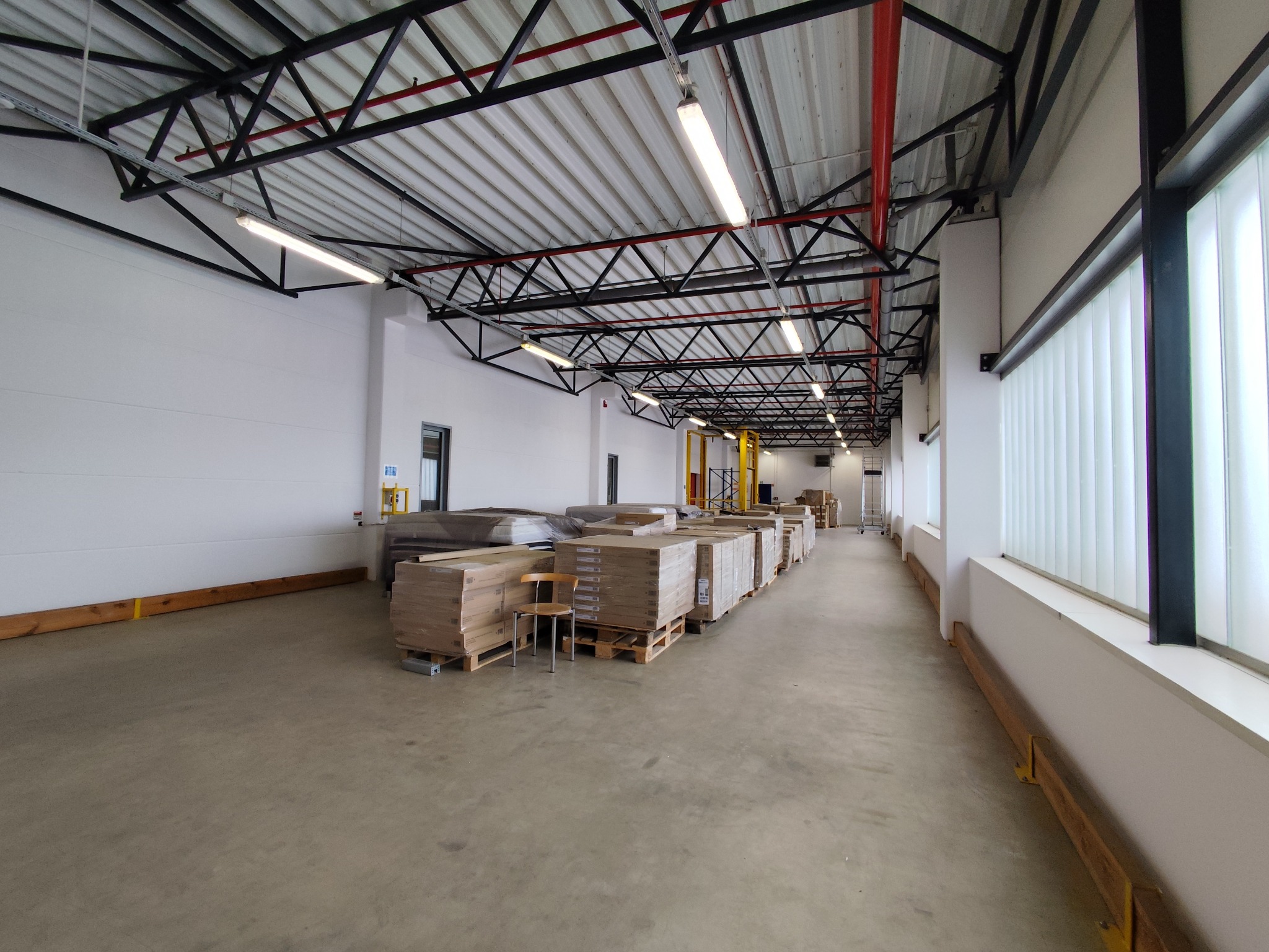 Magazyn z paletami płyt gipsowo-kartonowych i krzesłem. Widoczne białe ściany, metalowa konstrukcja dachu i okna z białymi lamelami. Perspektywa z głębi magazynu.