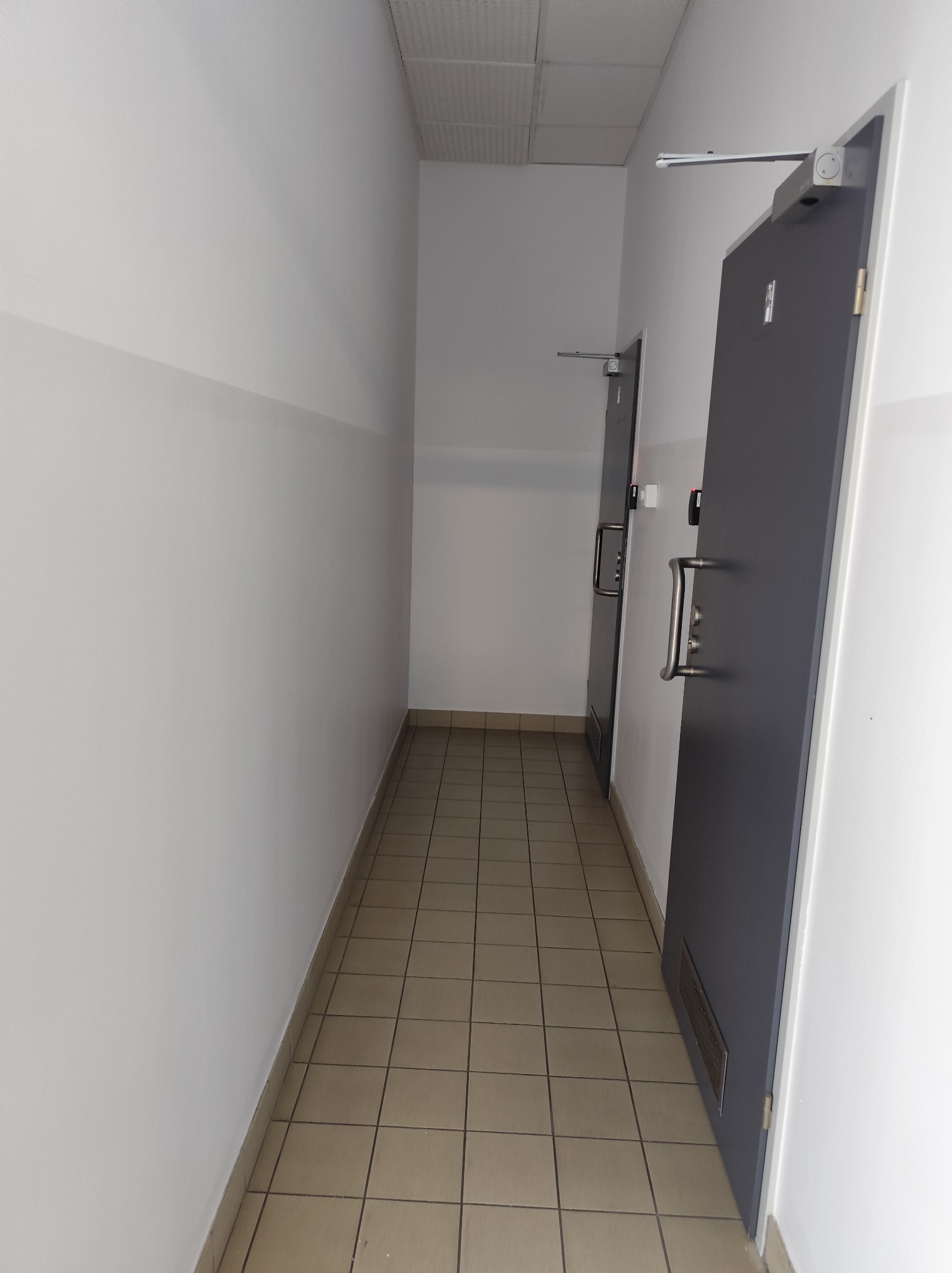Korytarz z dwiema szarymi drzwiami z metalowymi klamkami i systemem samozamykającym. Podłoga wyłożona kwadratowymi płytkami w odcieniach beżu. Ściany białe z szarym pasem.