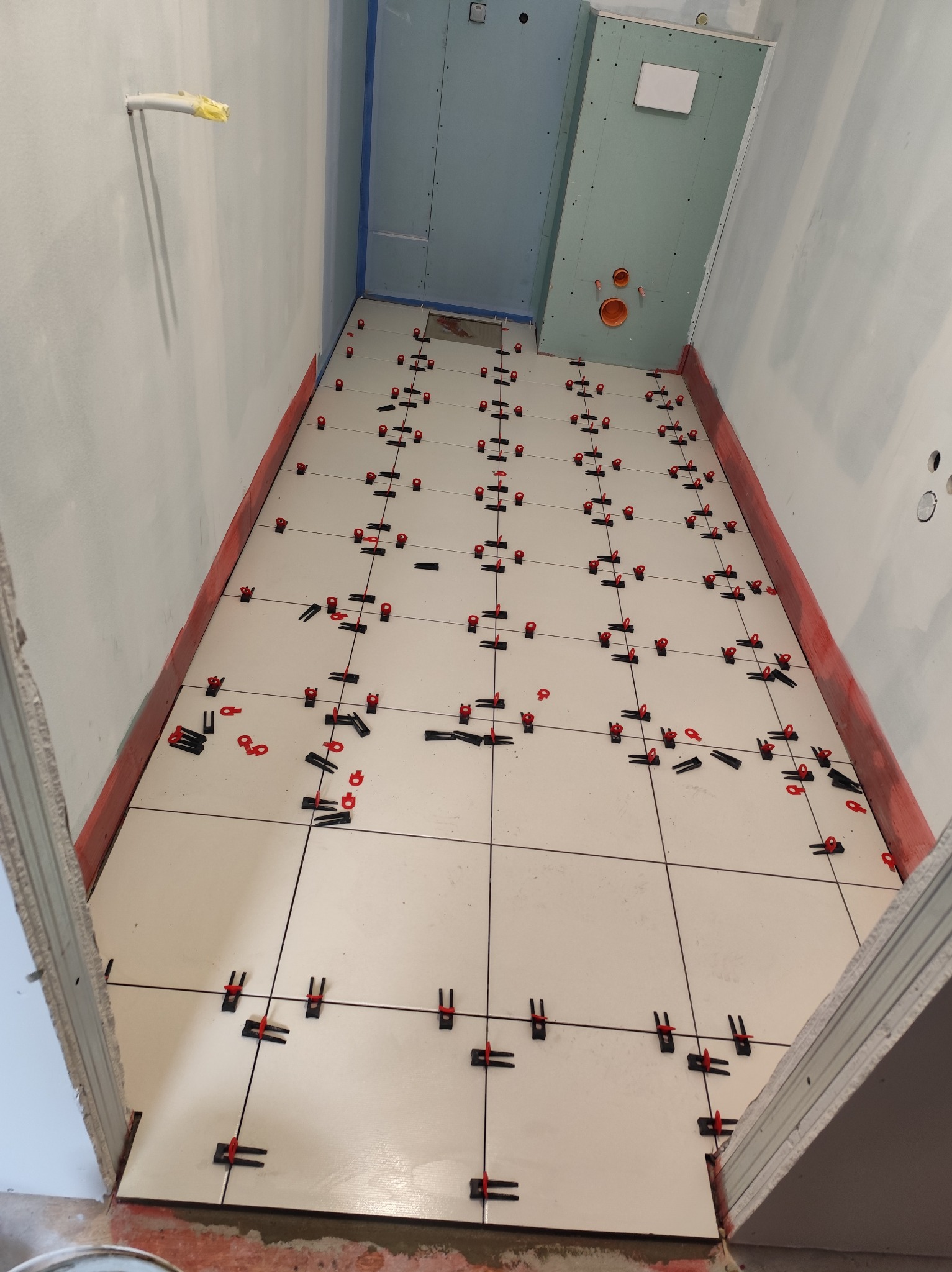 Białe płytki podłogowe w trakcie układania w wąskiej łazience. Czerwone klipsy poziomujące. Ściany przygotowane pod płytki, częściowo z płyt karton-gips.