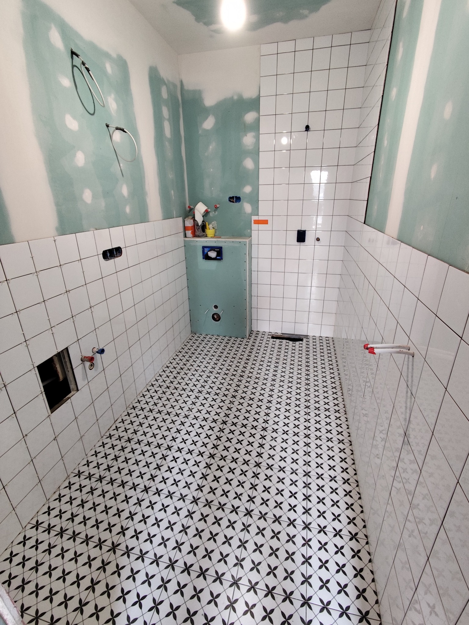 Łazienka w trakcie wykończenia: białe płytki na ścianach, wzorzysta podłoga, zabudowa stelaża WC. Widoczne instalacje i płyty gipsowo-kartonowe.