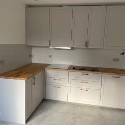 MW-Meble Wiśniewski - Jasnoszara zabudowa kuchenna z drewnianym blatem i miedzianymi uchwytami. Minimalistyczny styl, płytki w tle, indukcja. Ujęcie szerokokątne, widok na całą zabudowę.