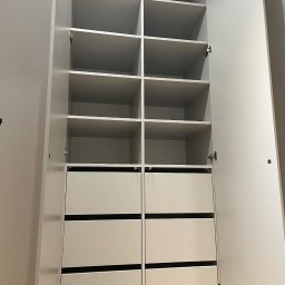 MW-Meble Wiśniewski - Otwarta, biała szafa z półkami i szufladami z czarnymi uchwytami, widok z dołu. Minimalistyczny design, jasne wnętrze, drzwi szafy otwarte na oścież.