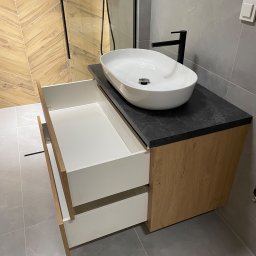 MW-Meble Wiśniewski - Nowoczesna szafka łazienkowa z dwiema otwartymi szufladami, blatem z czarnego kamienia i owalną umywalką. W tle płytki w jodełkę i szklana kabina prysznicowa.
