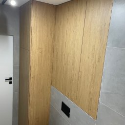 MW-Meble Wiśniewski - Drewniana zabudowa ściany z ukrytą szafą i toaletą w nowoczesnej łazience z szarą płytką. Minimalistyczny design z oświetleniem punktowym.