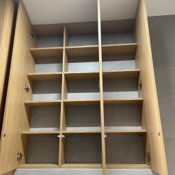 MW-Meble Wiśniewski - Otwarta, drewniana szafka z półkami, widok z dołu. Jasne drewno, szare płytki w tle. Szafka bez zawartości, z widocznymi zawiasami.