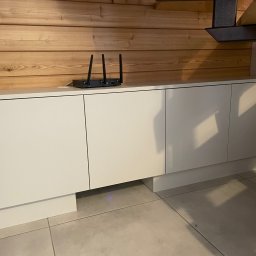 MW-Meble Wiśniewski - Białe szafki kuchenne z drewnianym panelem nad blatem, na którym stoi router. Fragment okapu widoczny w prawym górnym rogu.