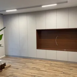 Jasnoszara szafa wnękowa z drewnianą wnęką na TV i półkami, oświetlenie LED na suficie, parkiet w jodełkę. Elementy wykończeniowe w stylu klasycznym.