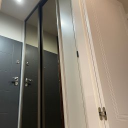 MW-Meble Wiśniewski - Nowoczesna szafa wnękowa z lustrzanymi drzwiami przesuwnymi, widoczna obok biała ściana z drzwiami. Ujęcie z dołu, ukazujące montaż i wykończenie.