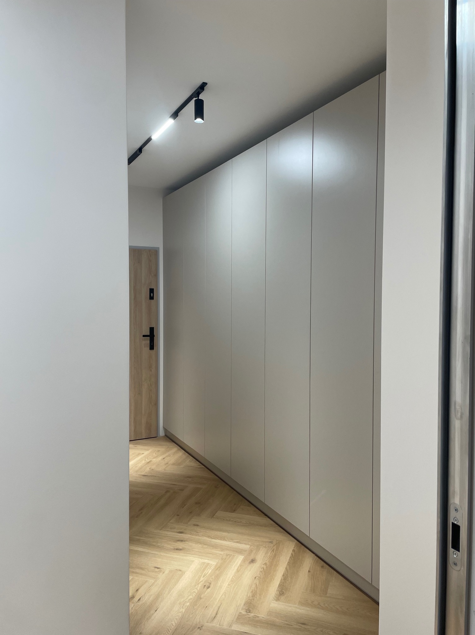 Jasna szafa wnękowa z gładkimi frontami, oświetlenie punktowe, parkiet w jodełkę. Minimalistyczny design, eleganckie wykończenie wnętrza, widok z perspektywy korytarza.