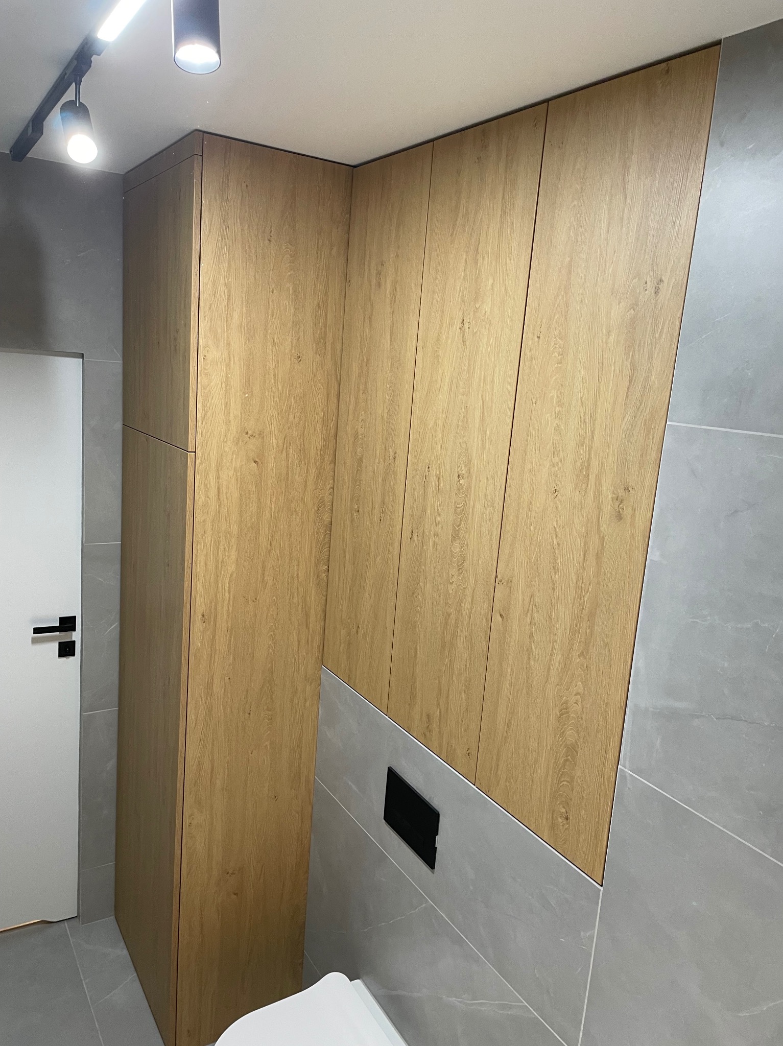 Drewniana zabudowa ściany z ukrytą szafą i toaletą w nowoczesnej łazience z szarą płytką. Minimalistyczny design z oświetleniem punktowym.