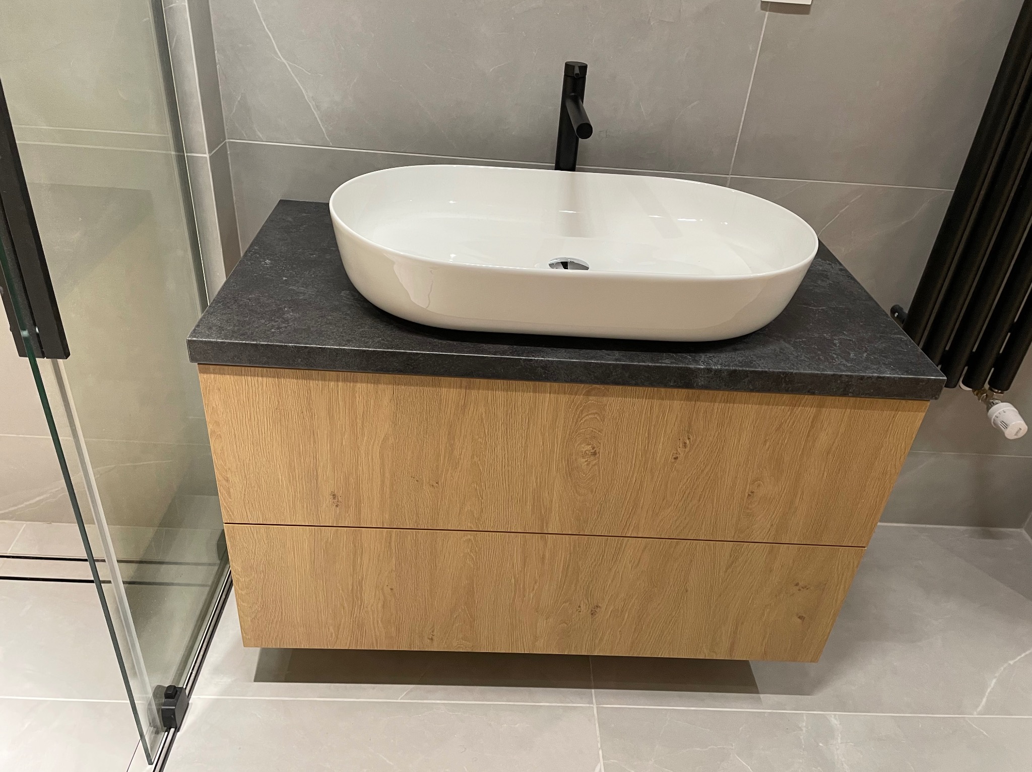 Nowoczesna szafka łazienkowa z drewnianymi frontami i owalną umywalką nablatową, czarna armatura, szare płytki w tle.