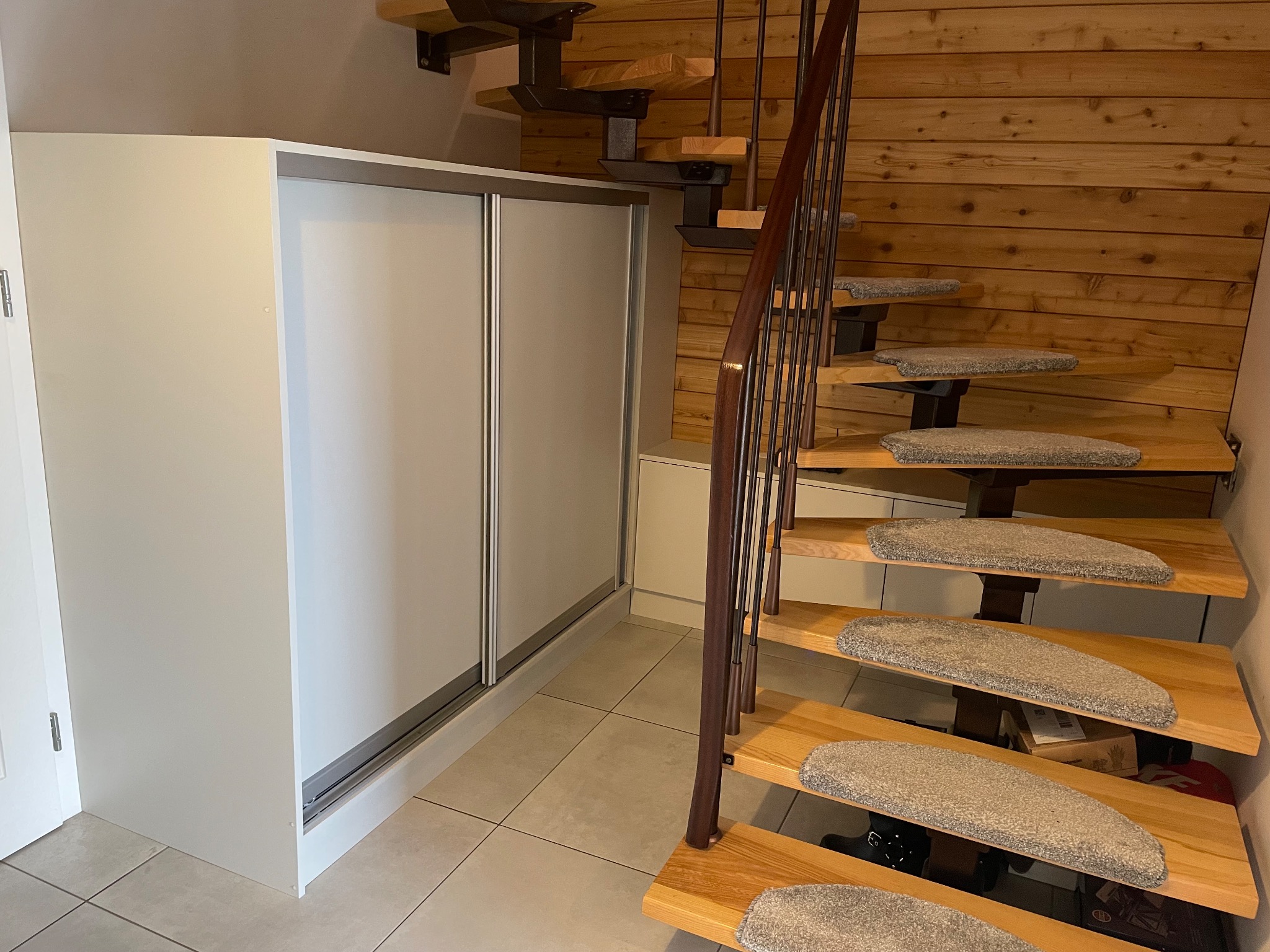 Biała szafa przesuwna obok drewnianych schodów z metalową balustradą i dywanikami na stopniach, na tle drewnianej ściany.