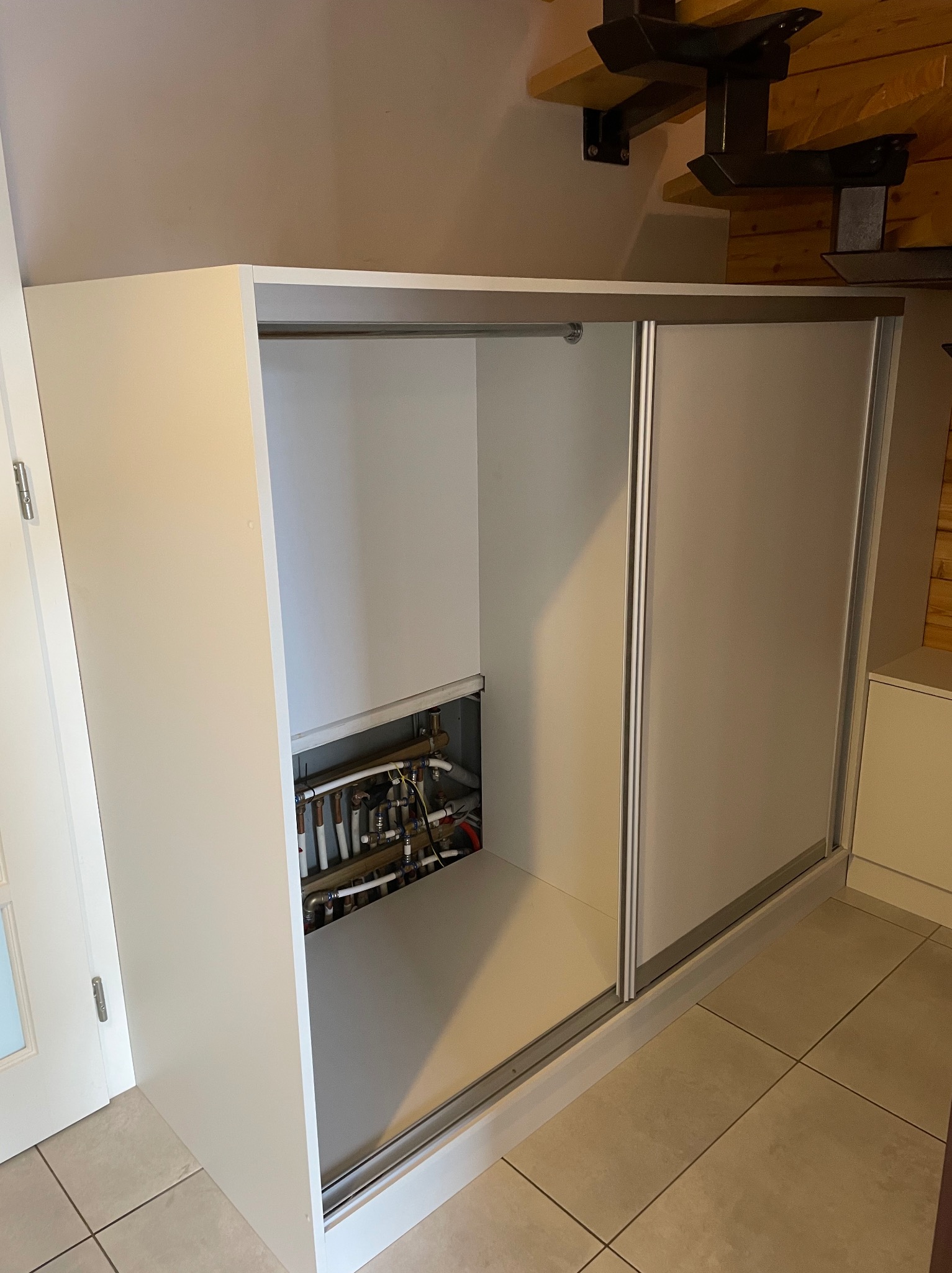 Biała szafa wnękowa z przesuwnymi drzwiami, częściowo odsłaniająca instalację grzewczą. Minimalistyczny design, jasne wnętrze, widoczne rury i złączki.