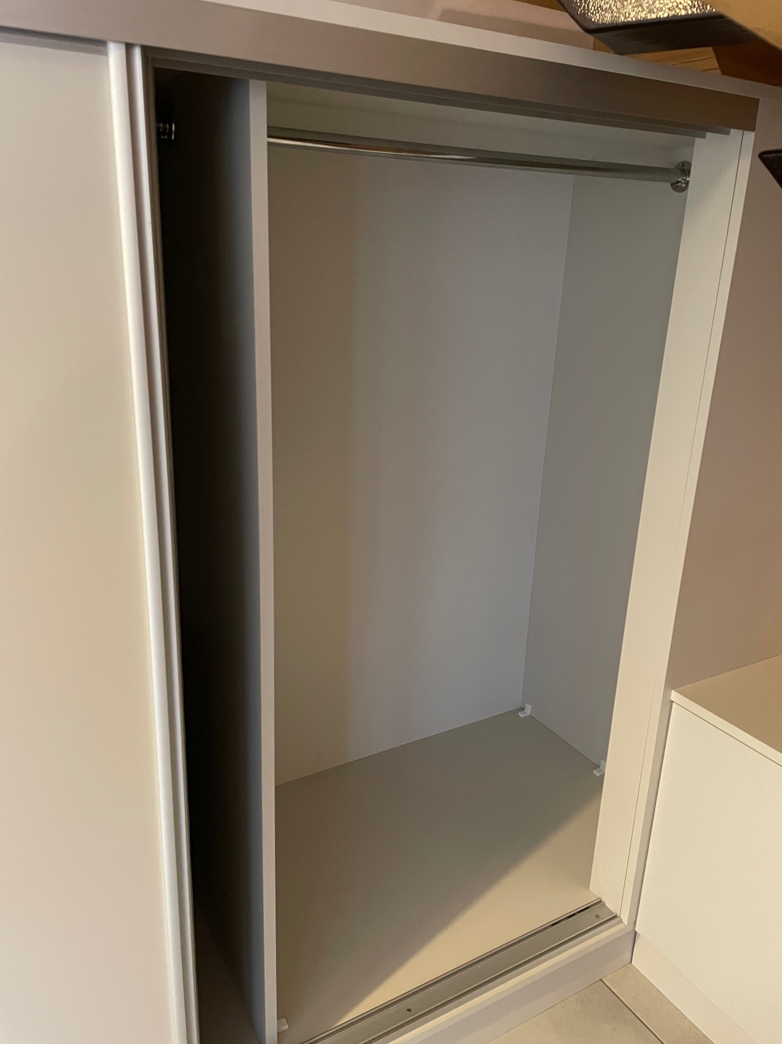 Wnętrze szafy wnękowej z drążkiem na ubrania i przesuwnymi drzwiami w odcieniach bieli i szarości. Widoczna szyna prowadząca drzwi.