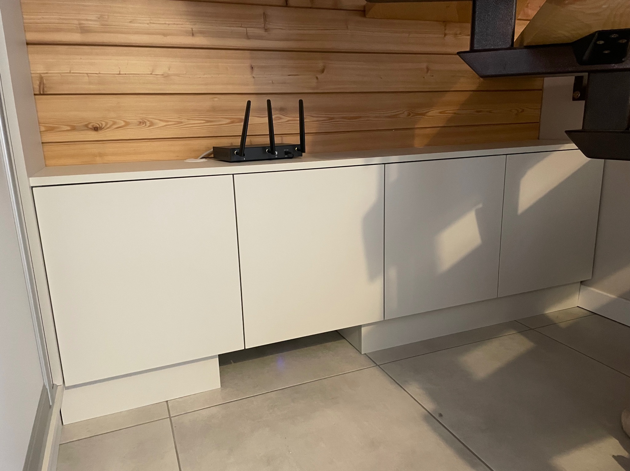 Białe szafki kuchenne z drewnianym panelem nad blatem, na którym stoi router. Fragment okapu widoczny w prawym górnym rogu.
