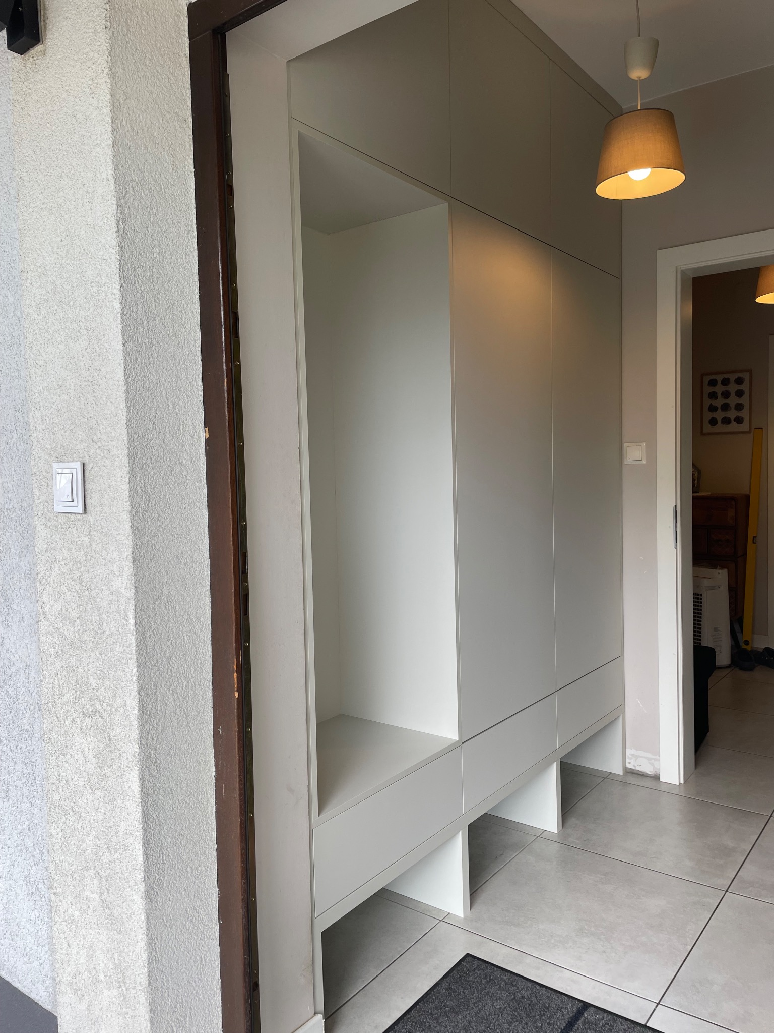 Biała szafa wnękowa z otwartą półką i szufladami, widoczna przy wejściu do domu, z szarą wycieraczką na podłodze z płytek.