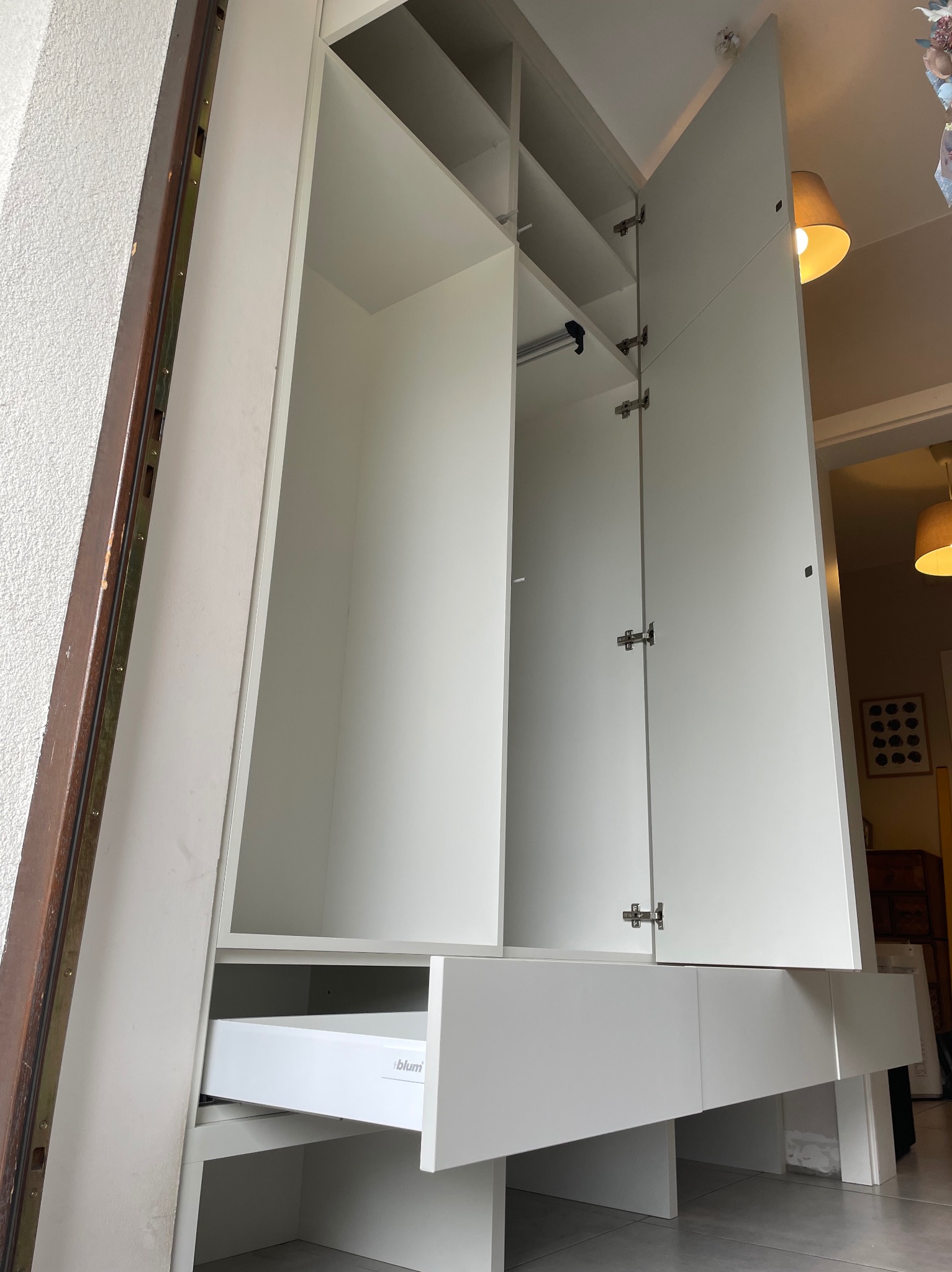 Biała szafa wnękowa z otwartymi drzwiami i wysuniętą szufladą Blum, widok z dołu, oświetlenie wnętrza, fragment ramy drzwiowej po lewej.