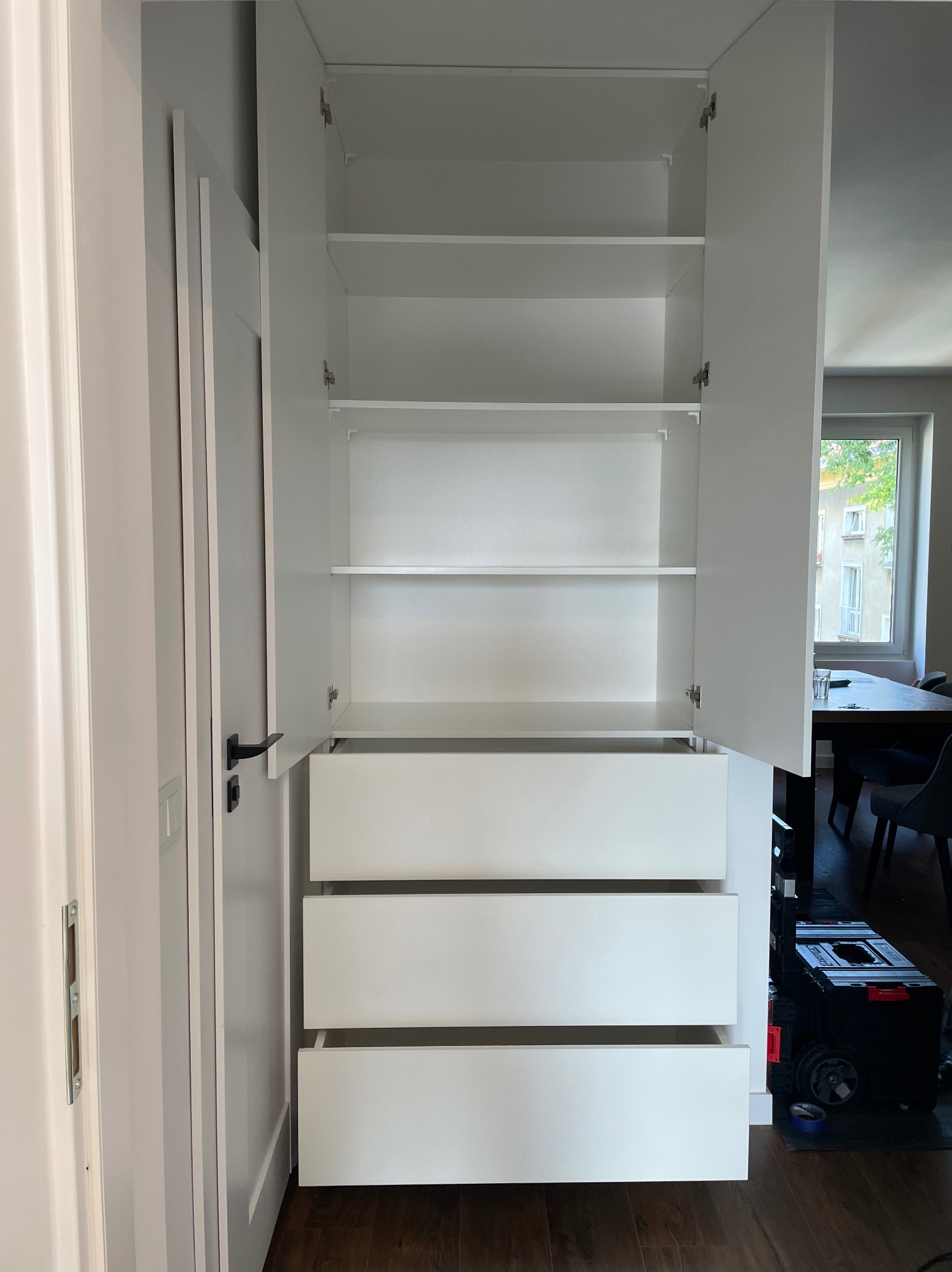 Biała szafa wnękowa z trzema szufladami i otwartymi półkami, widok z bliska na wykończone wnętrze i drzwi. W tle widoczne okno i stół.