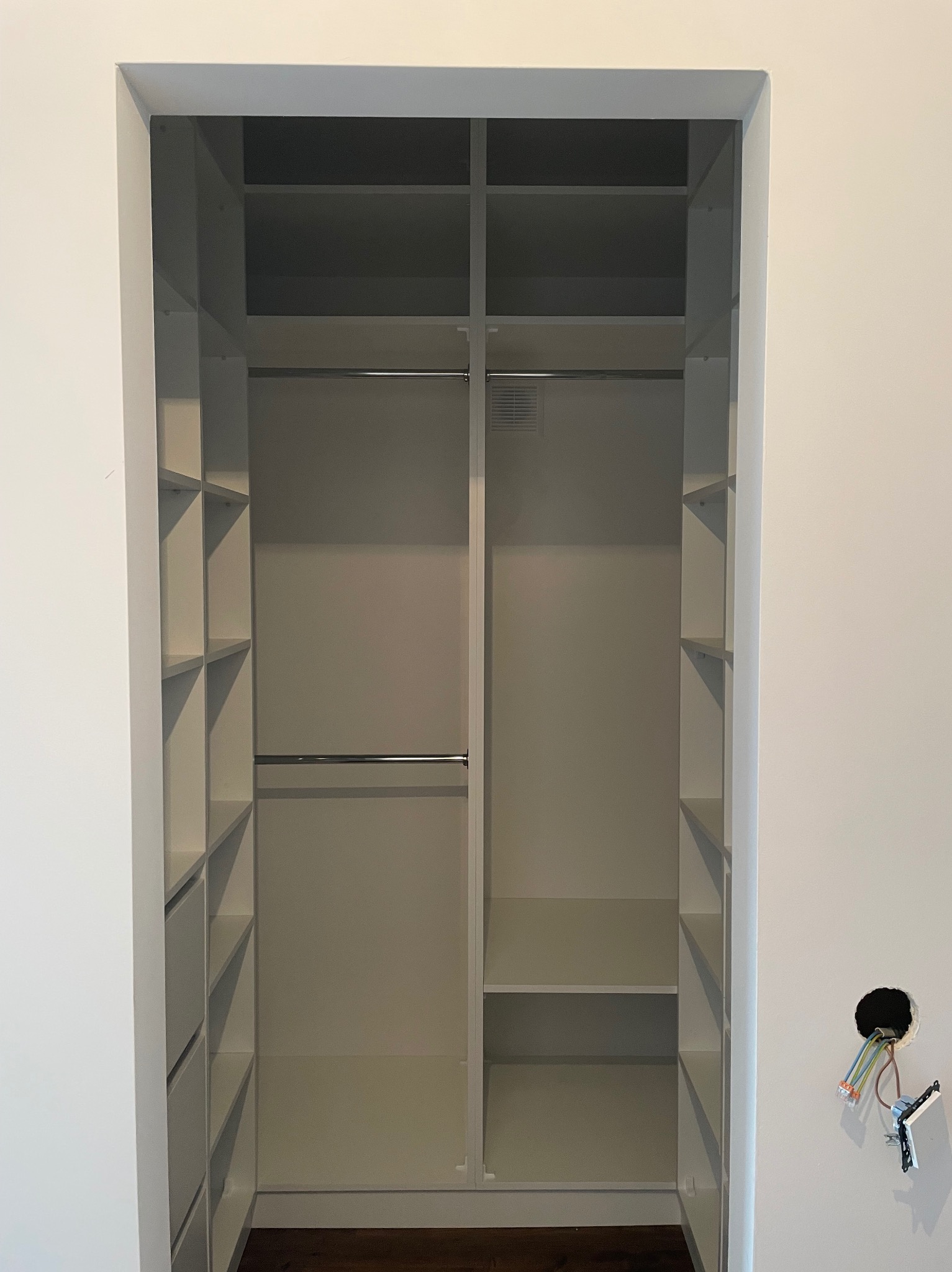 Wnętrze szafy wnękowej z półkami i drążkami na ubrania, wykończonej w bieli, w nowym budownictwie. Po prawej widoczne niedokończone gniazdko elektryczne.