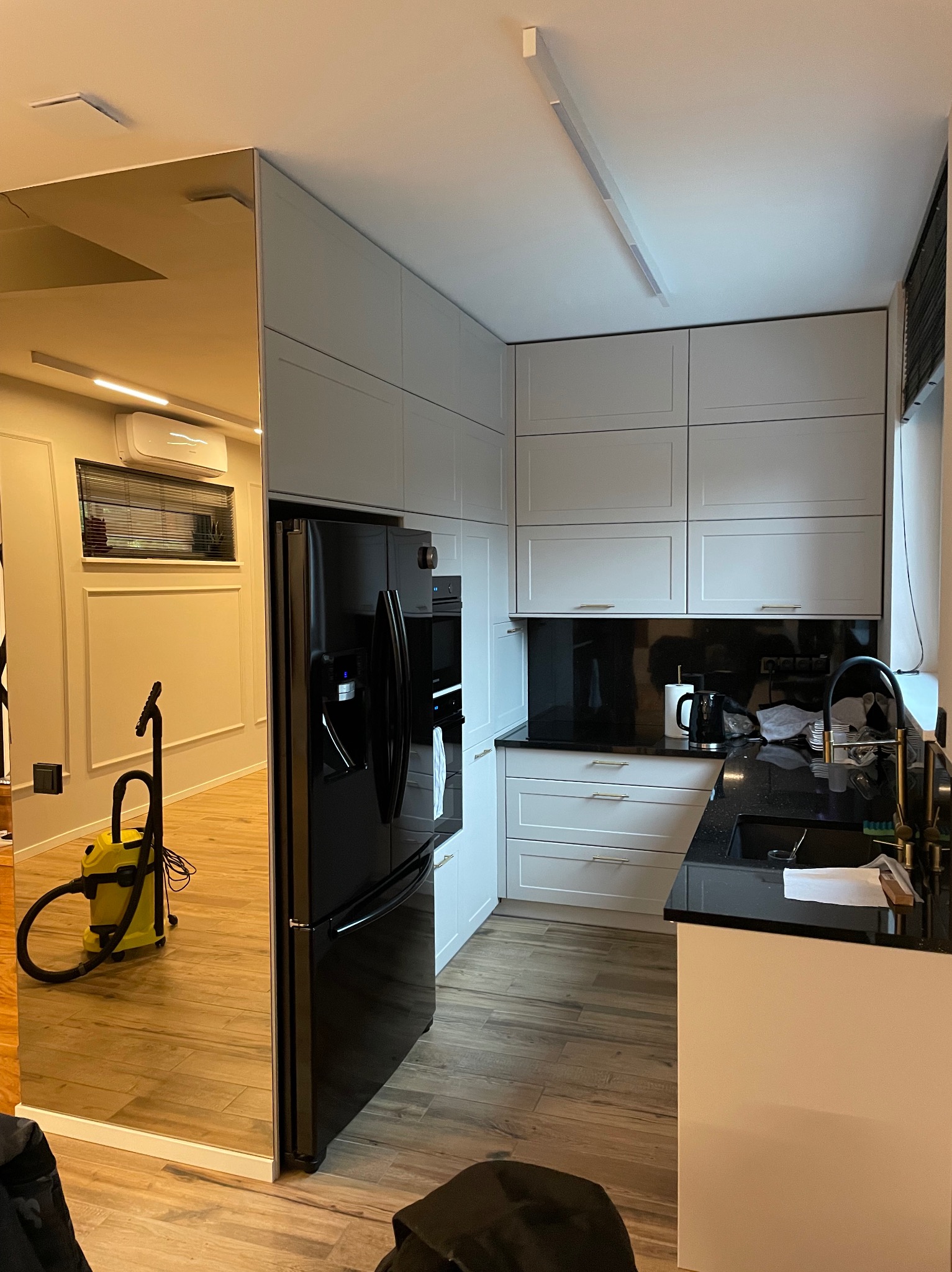 Nowoczesna kuchnia z białymi szafkami, czarnym blatem i lodówką. Lustrzana ściana odbija odkurzacz i wnętrze pokoju, tworząc iluzję większej przestrzeni.