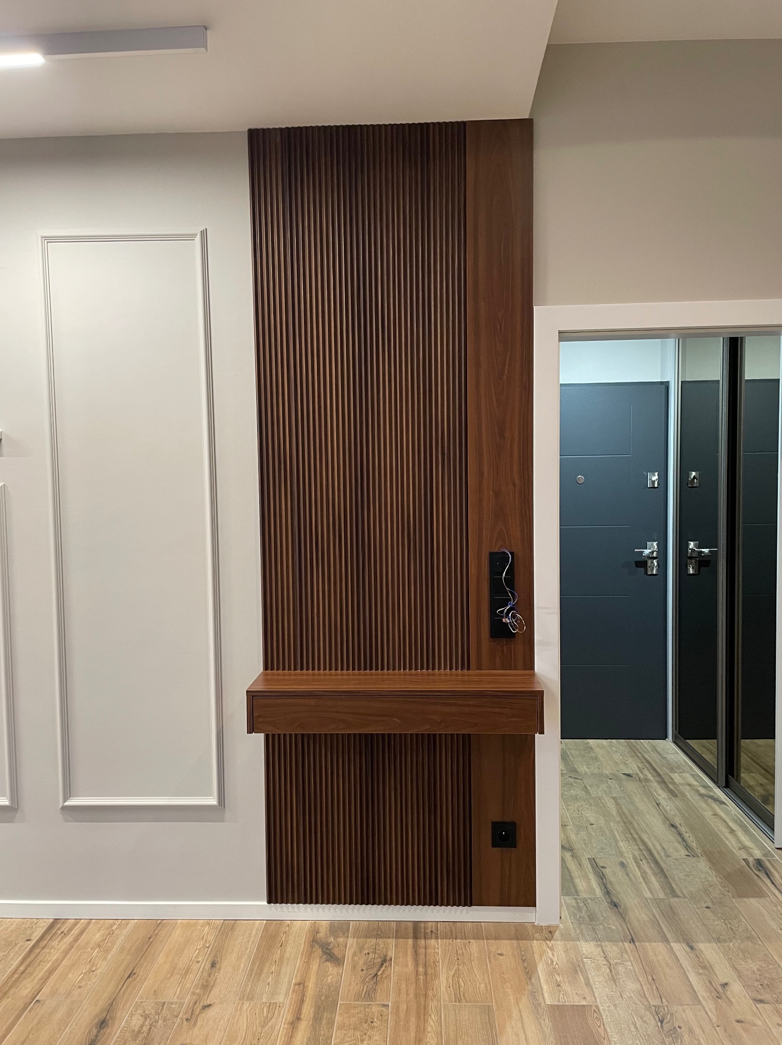 Nowoczesna szafka z drewnianymi lamelami i blatem z gniazdkiem, wkomponowana w przedpokój z panelami ściennymi i widokiem na drzwi.