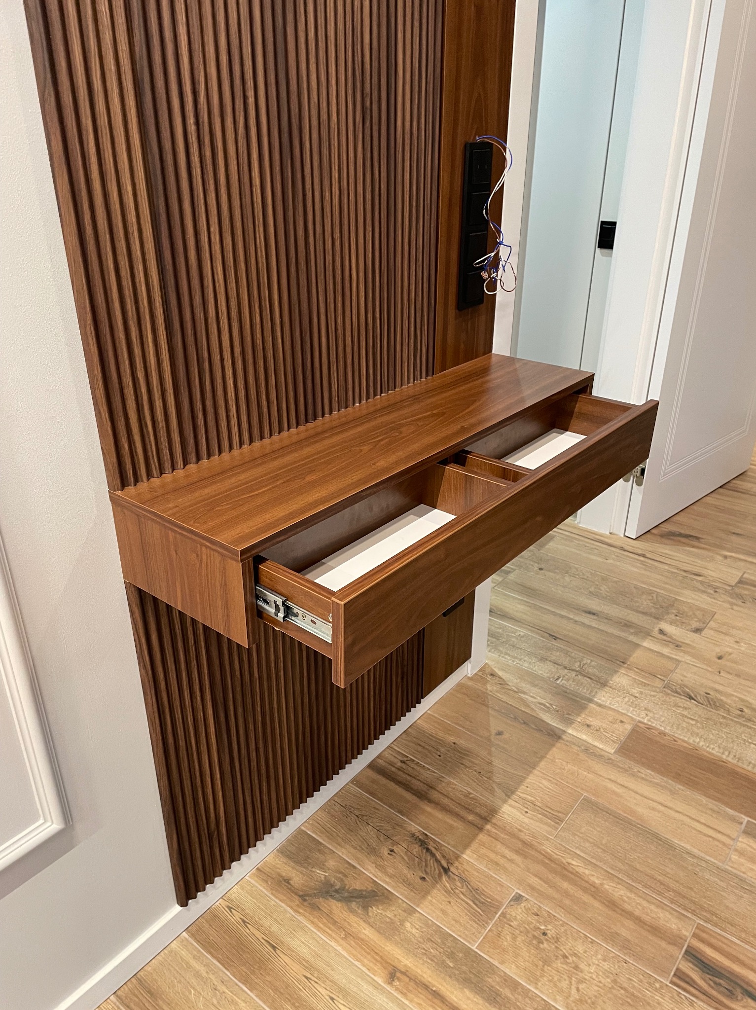 Drewniana szafka z wysuwaną szufladą, zamontowana na panelu ściennym z ryflowanego drewna. Widoczne gniazdka elektryczne i fragment podłogi z płytek imitujących drewno.