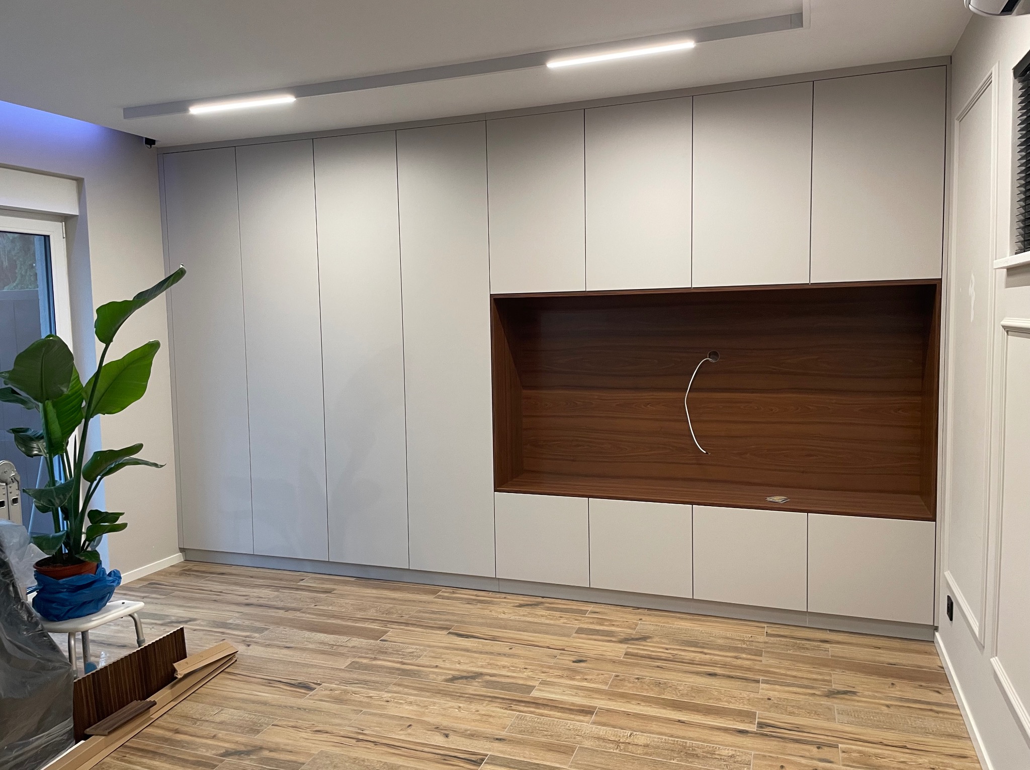 Jasnoszara szafa wnękowa z drewnianą wnęką na TV i półkami, oświetlenie LED na suficie, parkiet w jodełkę. Elementy wykończeniowe w stylu klasycznym.