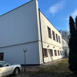 Dwupiętrowy budynek z białą elewacją i brązowymi ramami okiennymi, z widoczną drabiną opartą o ścianę, zaparkowany samochód i ozdobna latarnia uliczna z niebieską wstążką.