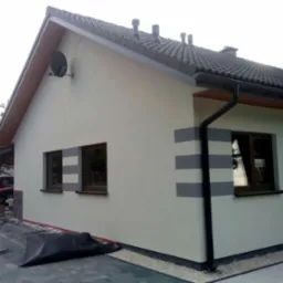 Ściana domu po tynkowaniu z poziomymi pasami szarej farby, widoczne okna z brązowymi ramami, dach z ciemnej dach&oacute;wki, przygotowane narzędzia i folia ochronna na ziemi.