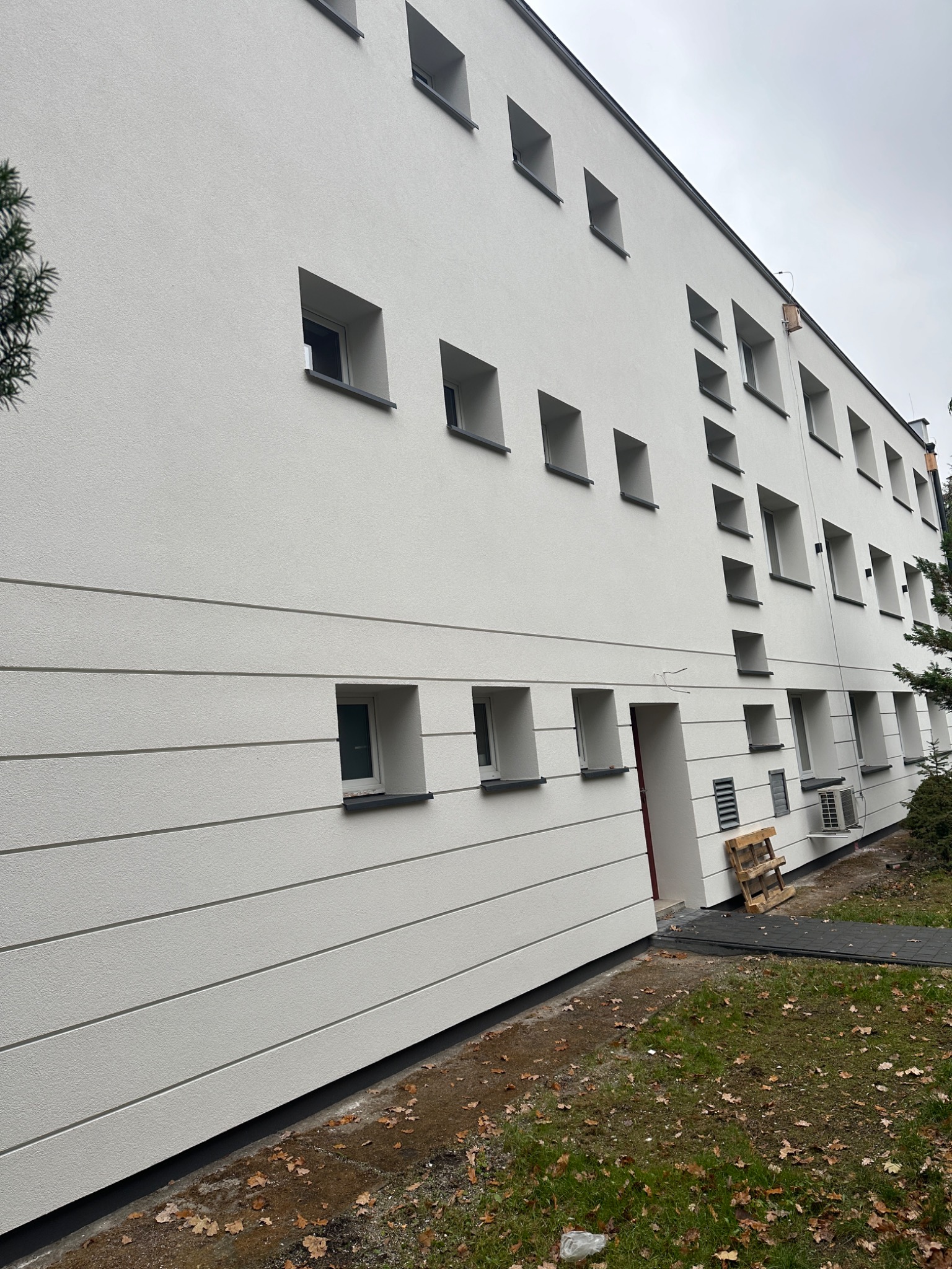 Nowoczesny budynek z białą elewacją o minimalistycznym designie, charakteryzujący się regularnym układem kwadratowych okien i poziomymi pasami dekoracyjnymi. Widoczny fragment terenu z trawą...