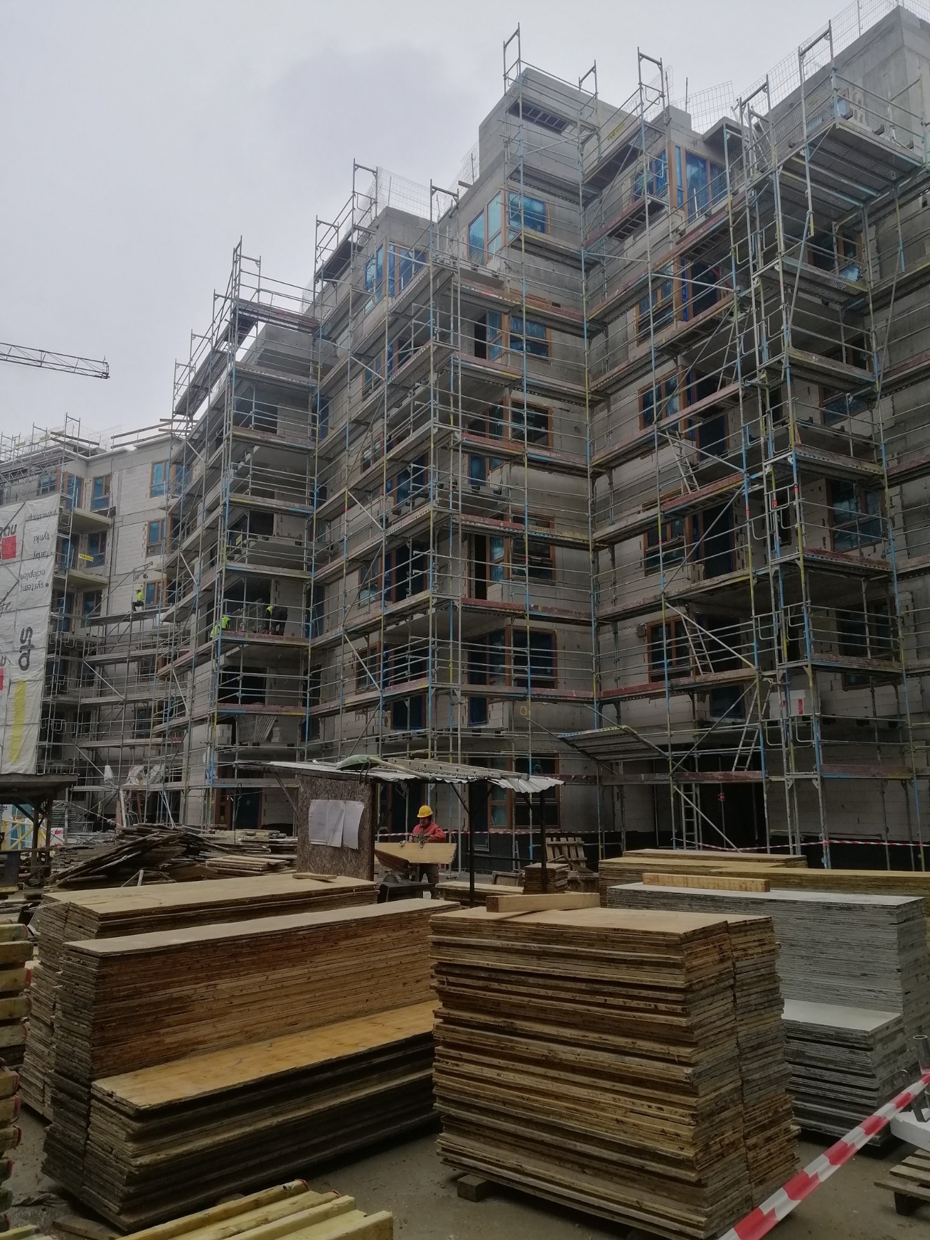 Wielopiętrowy budynek w trakcie budowy, otoczony rusztowaniami, z widocznymi oknami zabezpieczonymi folią i stertami drewnianych płyt na pierwszym planie, pracownik w kasku przenosi płytę.
