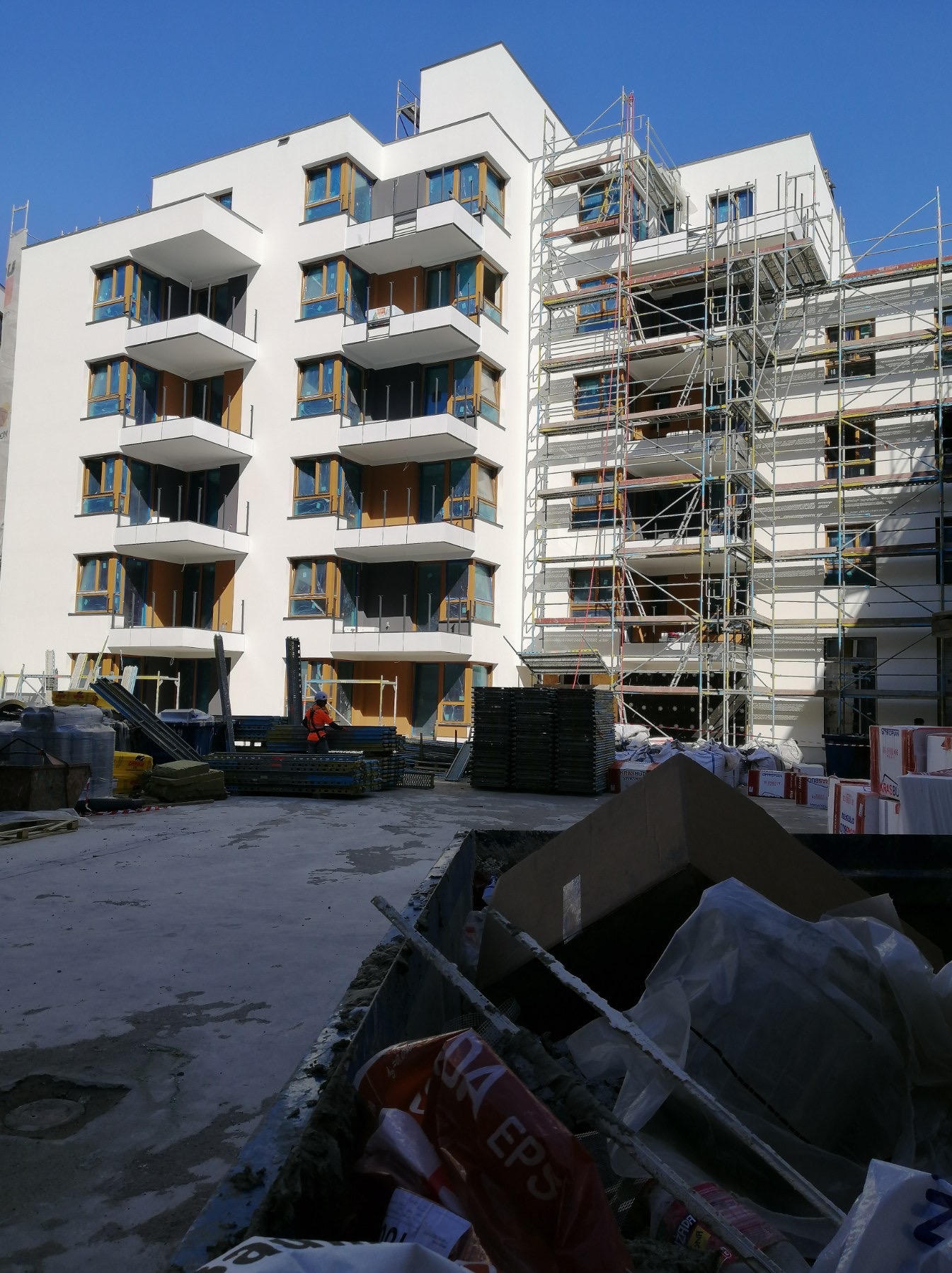 Nowoczesny, biały budynek wielorodzinny w trakcie prac elewacyjnych, z widocznymi rusztowaniami, materiałami budowlanymi i pracownikiem w pomarańczowej kamizelce, z perspektywy kontenera na odpady.