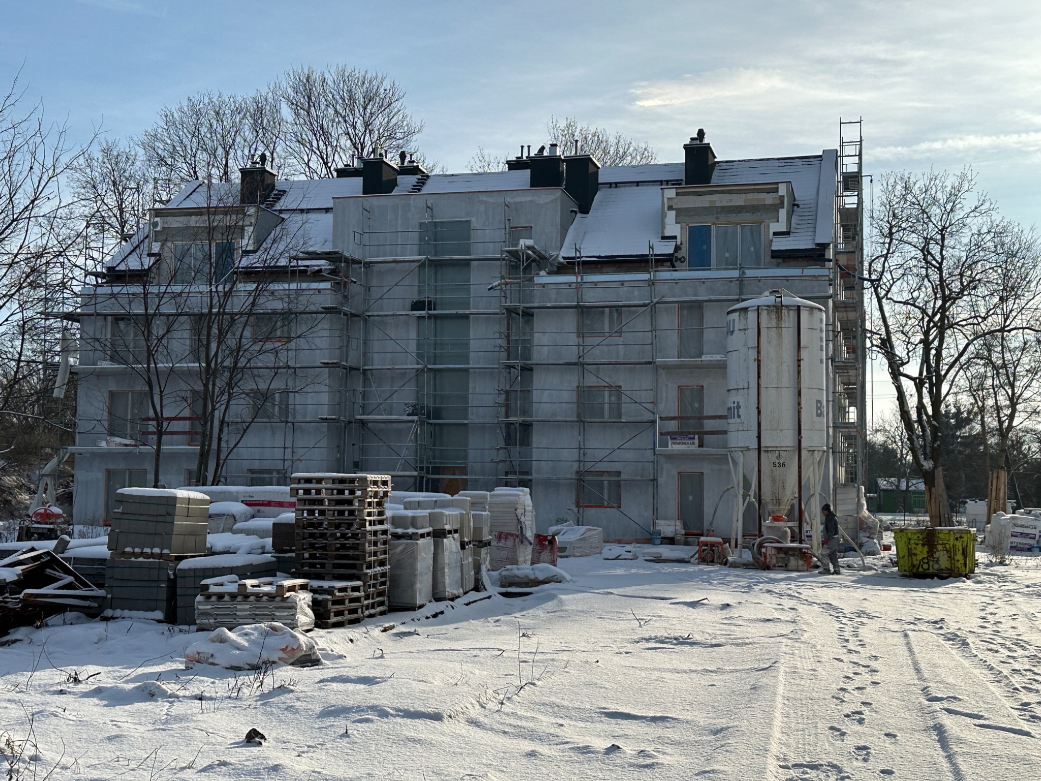 Wielopiętrowy budynek w trakcie prac elewacyjnych, osłonięty rusztowaniami i siatką, widoczny śnieg na dachu i ziemi, materiały budowlane składowane na placu budowy, pracownik w kombinezonie przy...